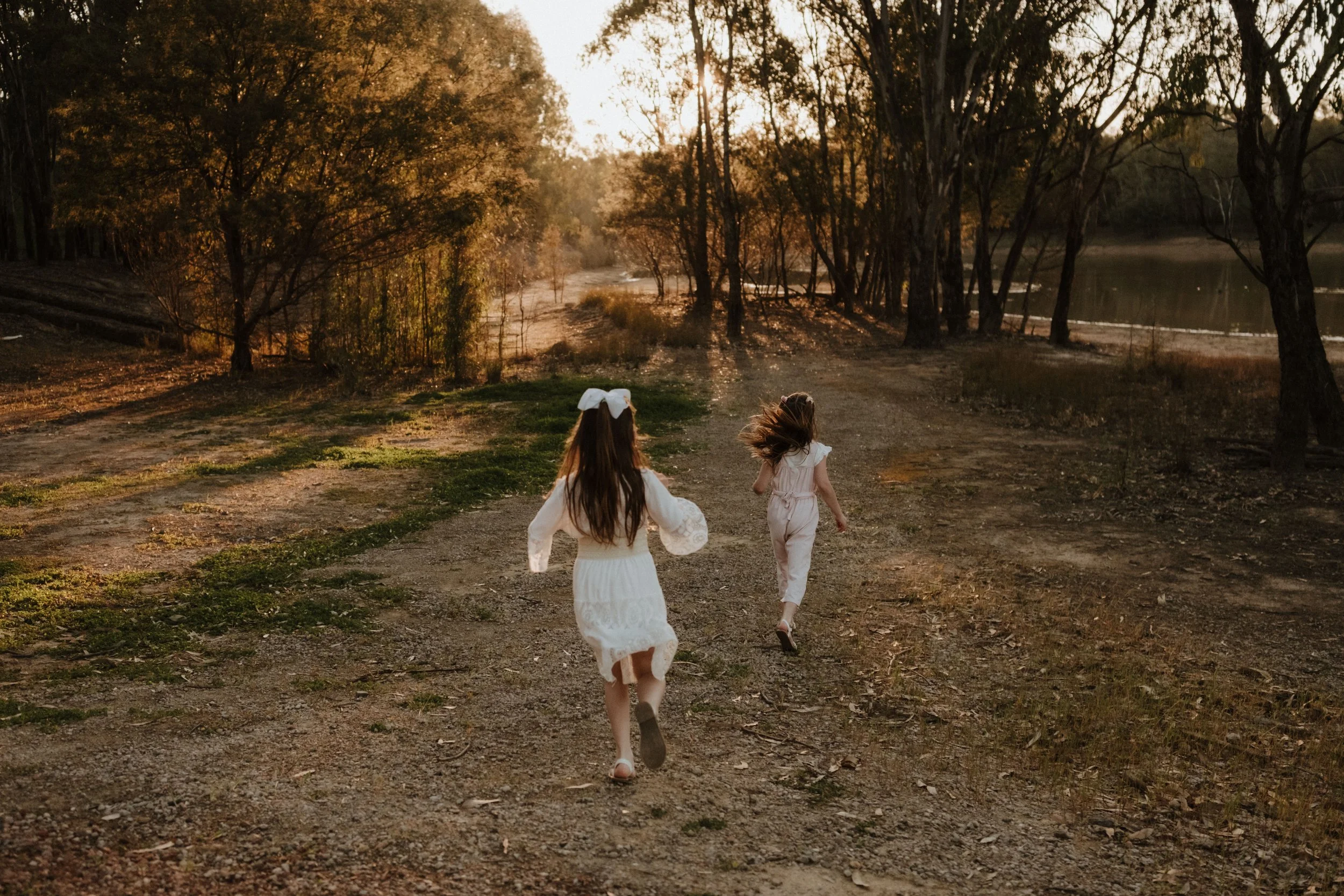 Two young girls running on a dirt trail near a lake at sunset, surrounded by trees with fall foliage.