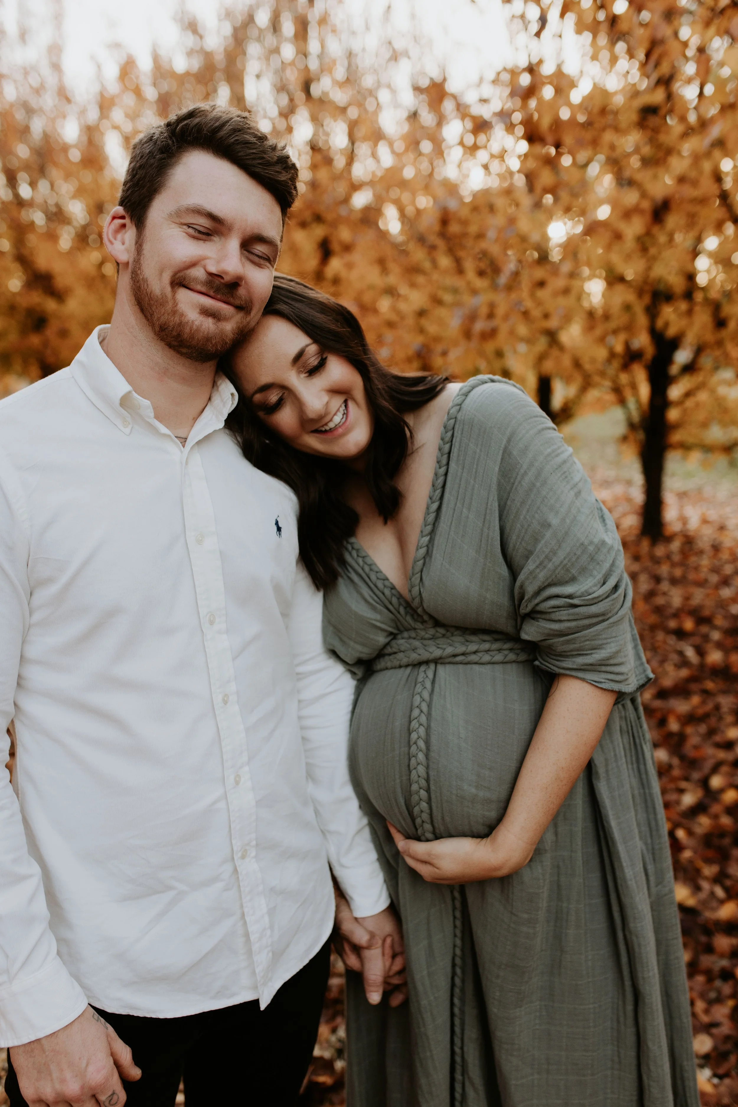 A pregnant woman and a man holding hands outdoors in fall, with trees and leaves in the background, the woman smiling- and resting her head on the man's shoulder.
