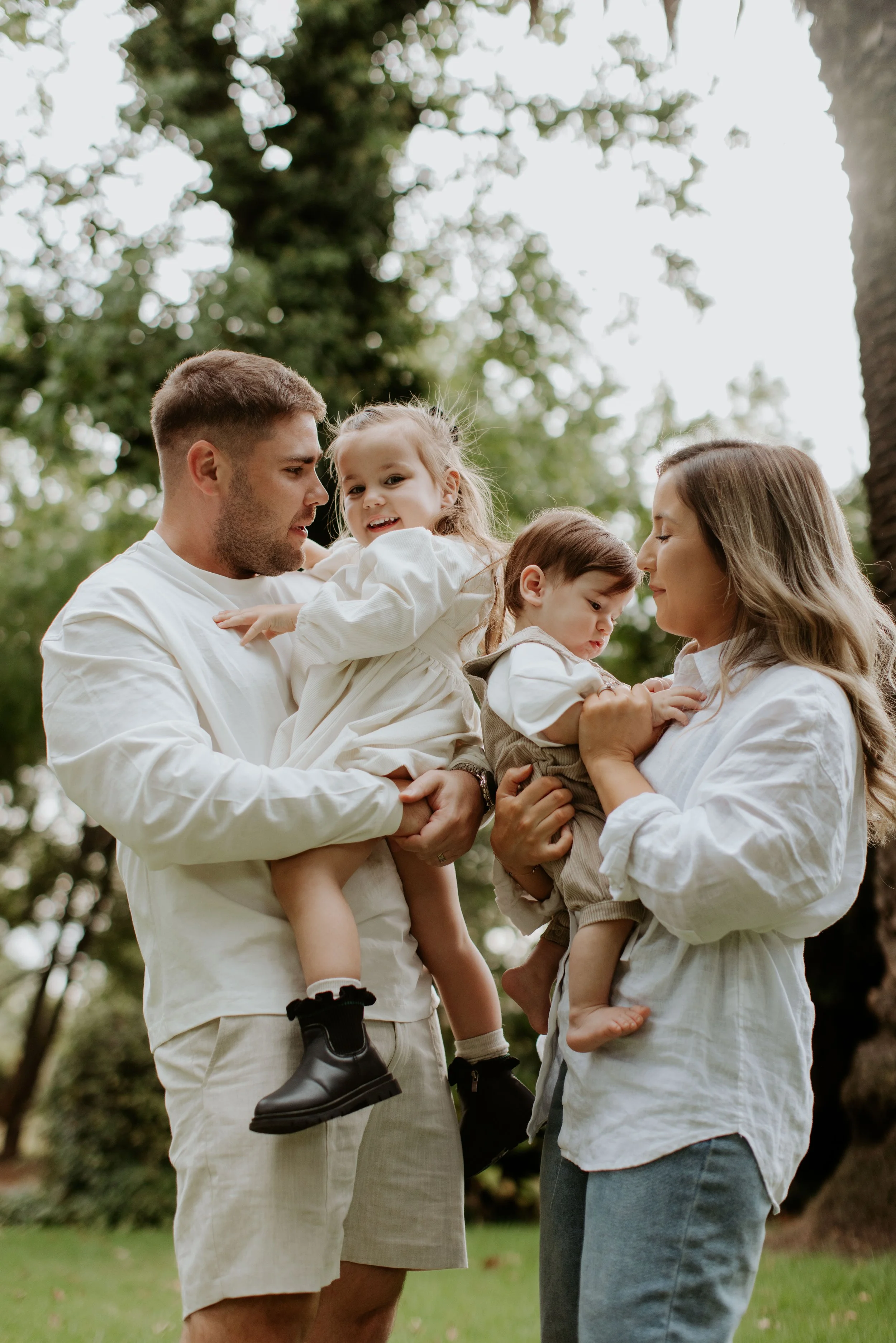 A family of four outdoors in a park, two adults and two children, with the children sitting on the adults' arms.