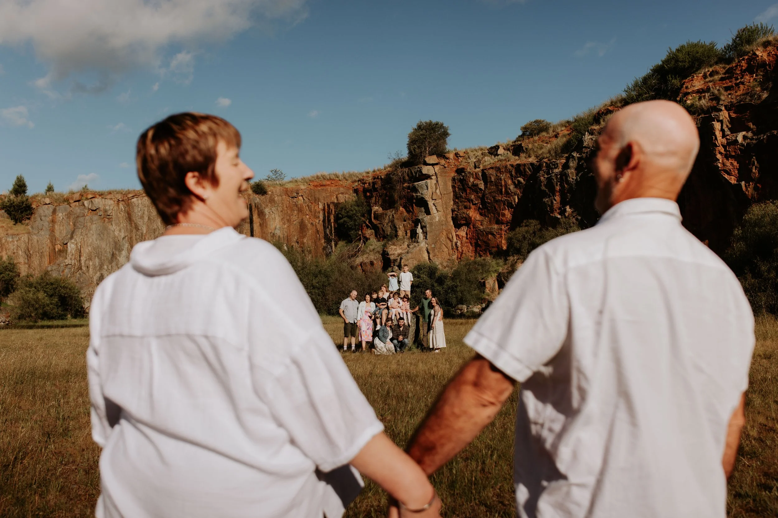 Two people holding hands in the foreground, looking at a group of people in the distance, set against a rocky hillside and clear sky.