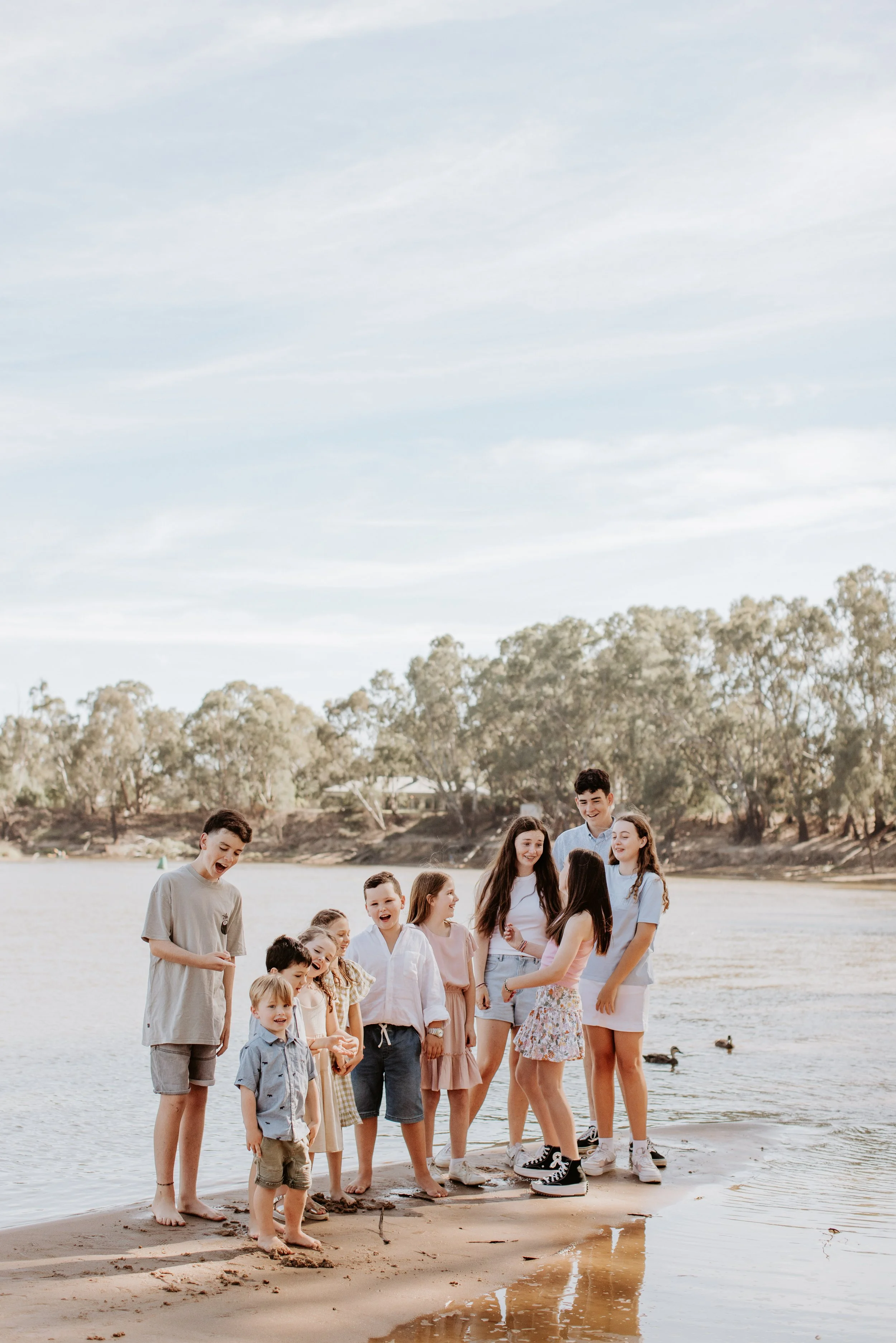 A group of children and teenagers standing by the water's edge on a sandy beach, laughing and chatting.