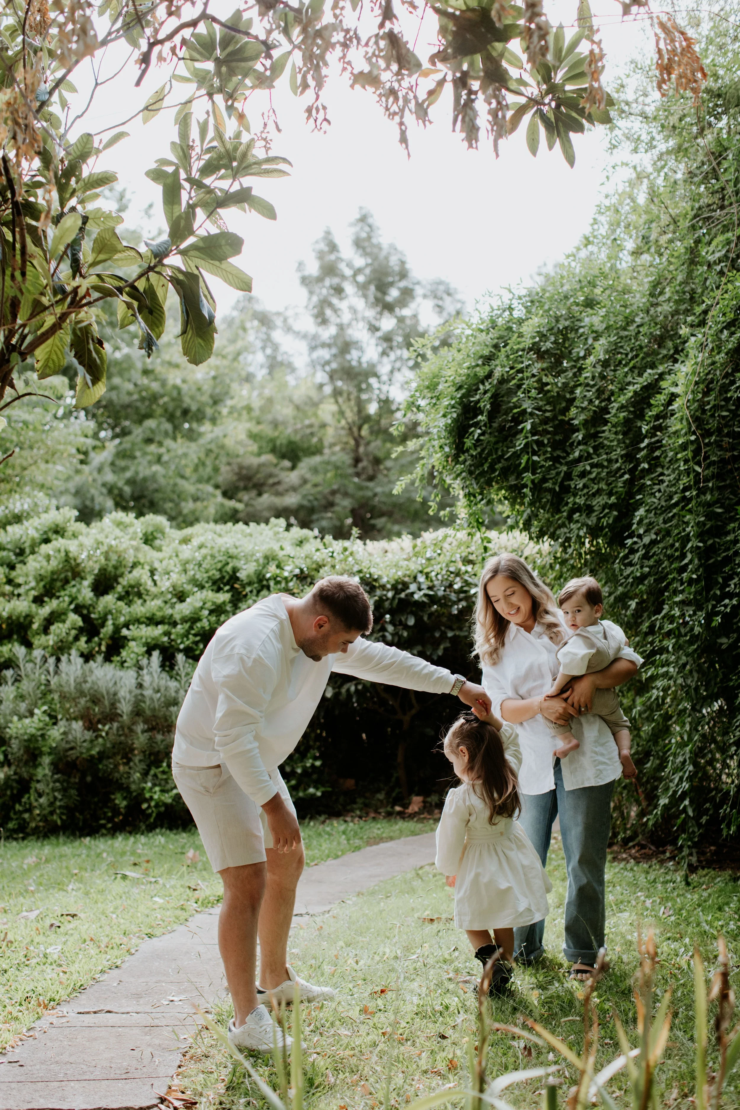 A family of four enjoying a day outdoors in a lush, green garden. The father is bending down to pet a young girl, while the mother is holding a toddler and smiling. The scene is bright and cheerful, with trees and bushes in the background.