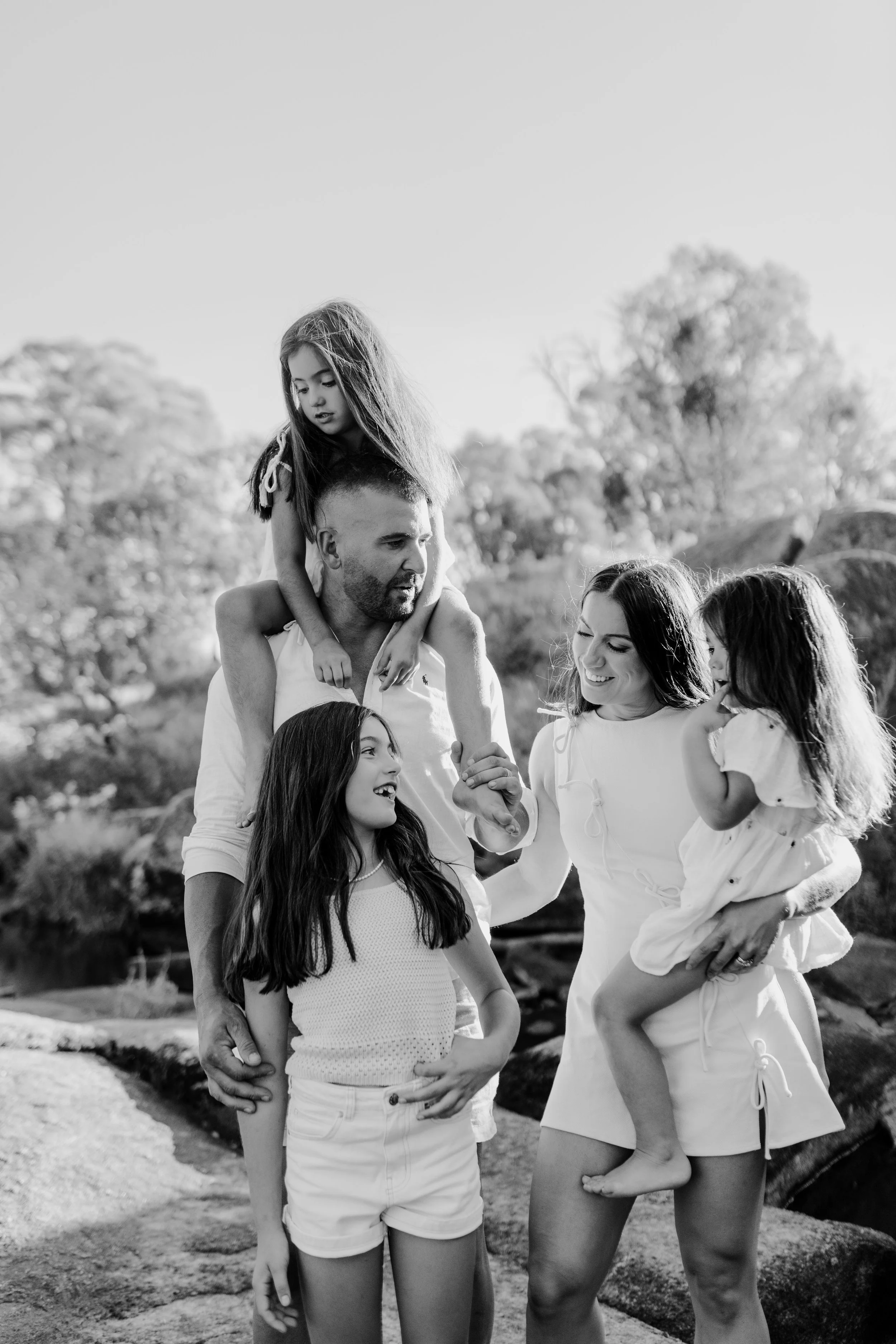 A happy family outdoor with two adults and three young girls, one girl on a man's shoulders, all smiling and holding hands, in black and white.