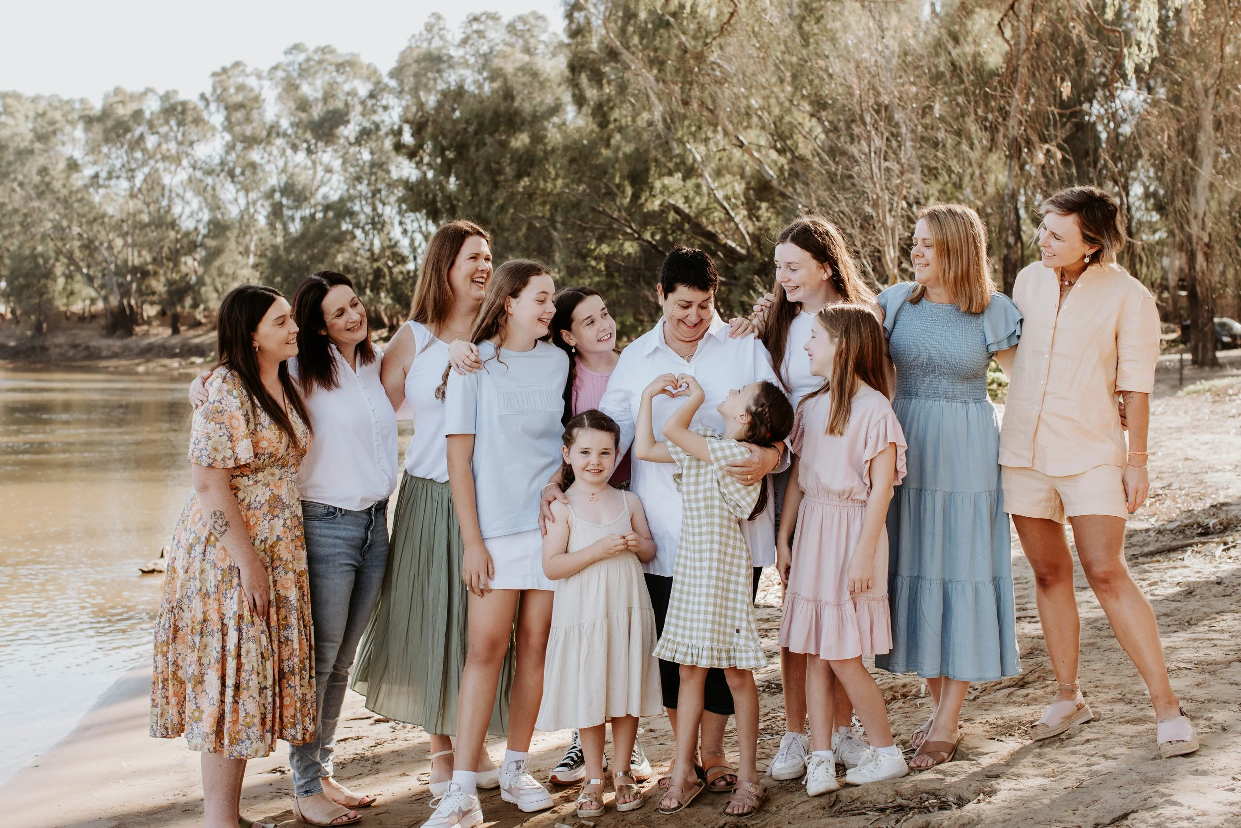 A diverse group of women and girls standing together on a sandy riverbank in a park, smiling and hugging in warm sunlight.