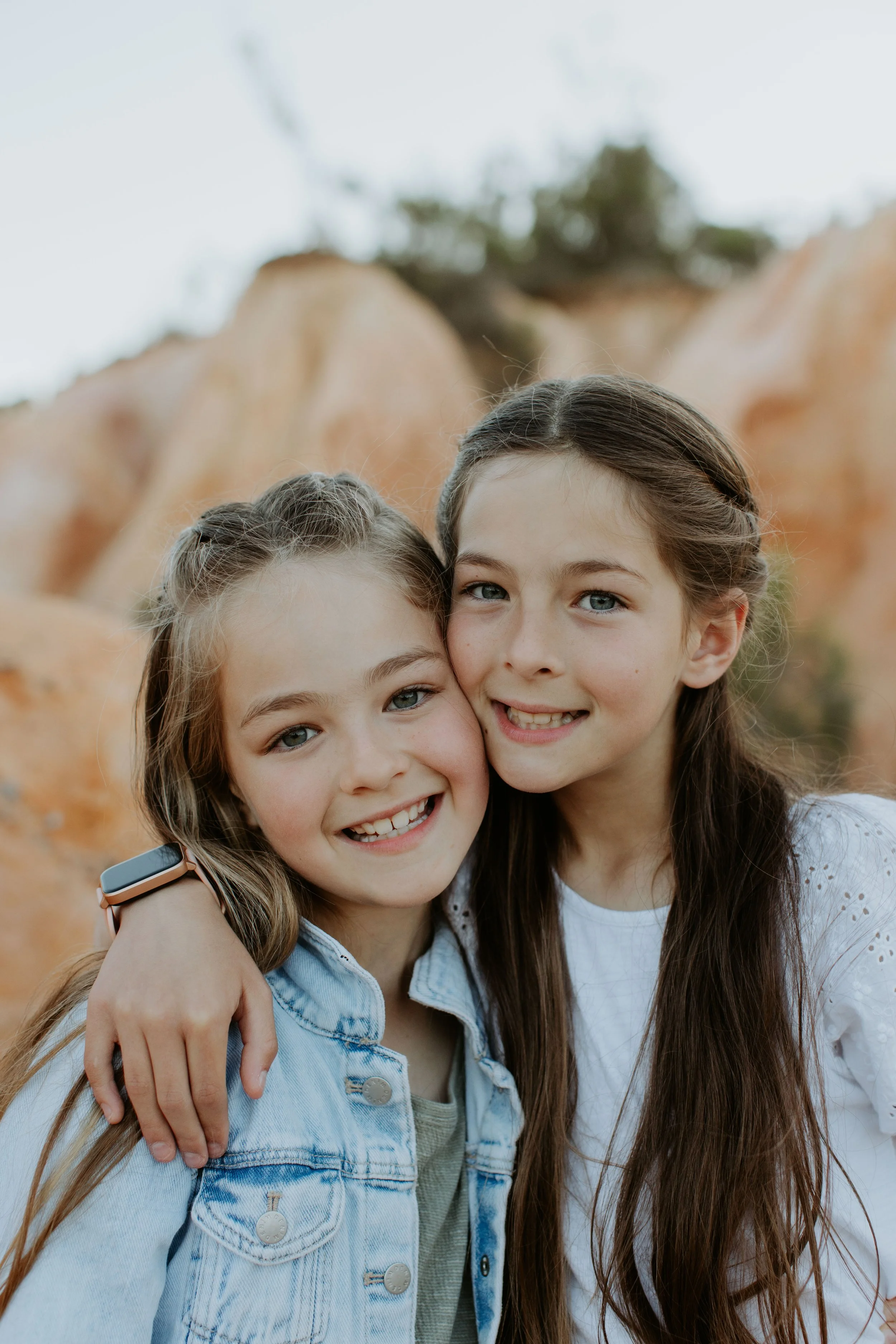 Two young girls smiling and hugging outdoors with rocky background and trees.
