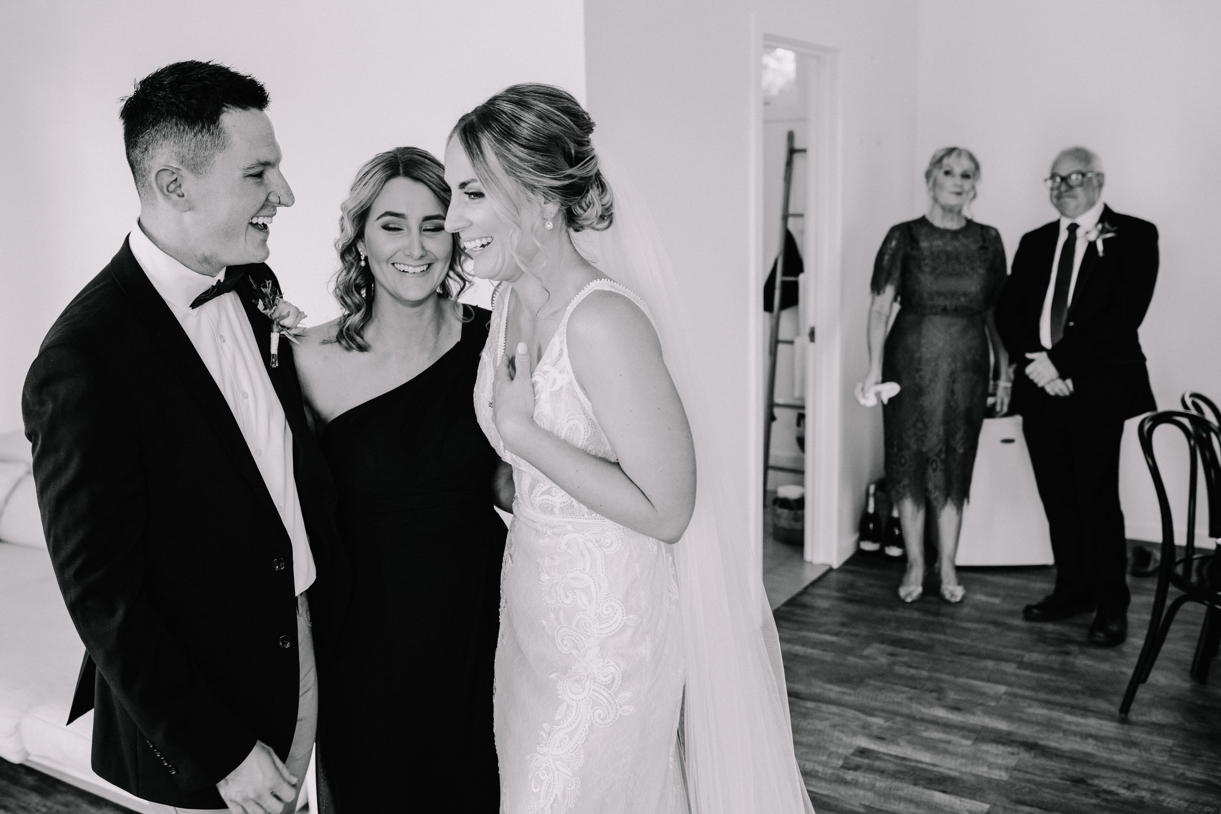 Black and white photo of a bride and groom smiling at each other, with a woman in a black dress between them, and two older adults in formal attire in the background, standing near a doorway.