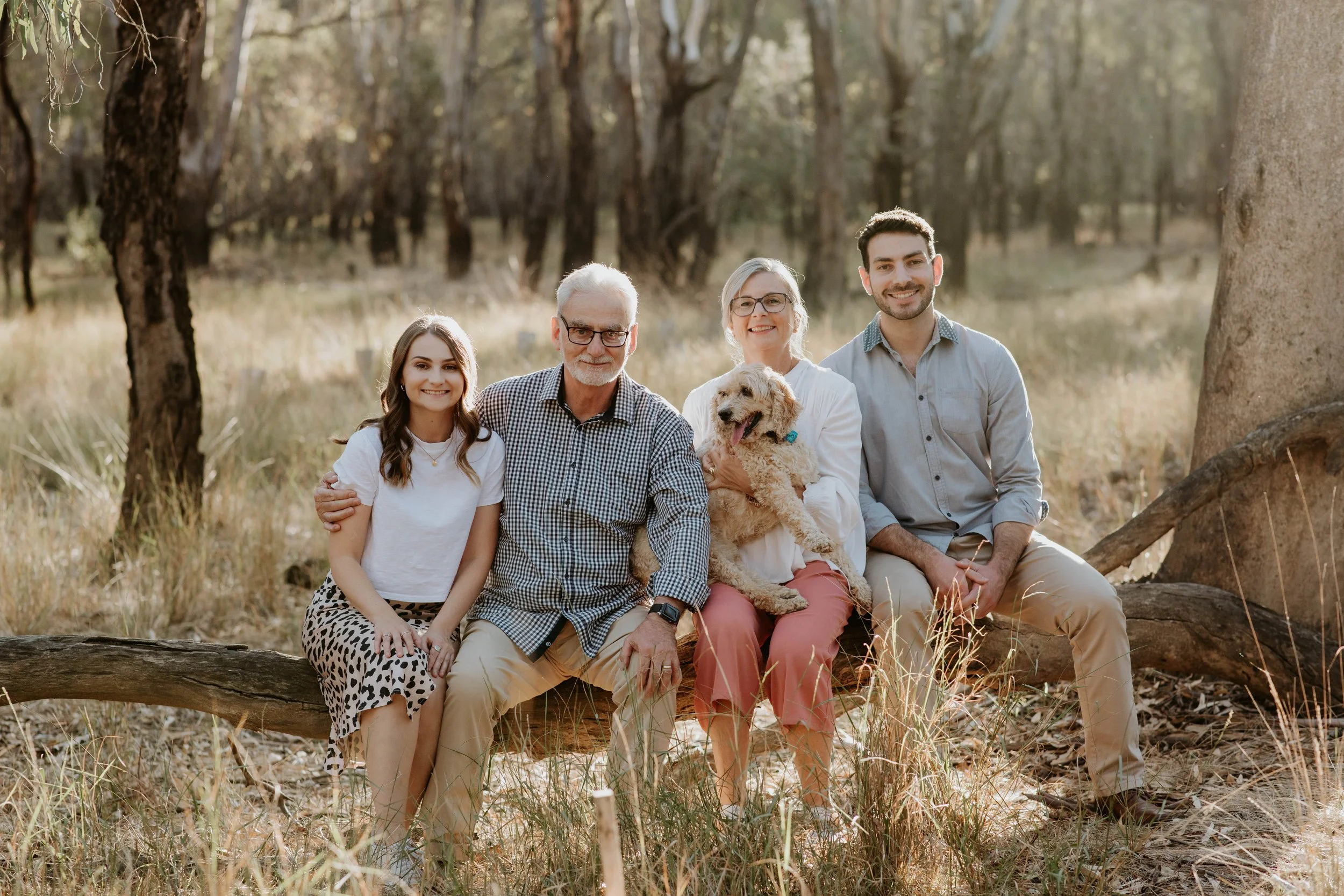 Family of five with a dog sitting outdoors on a log in a wooded area.