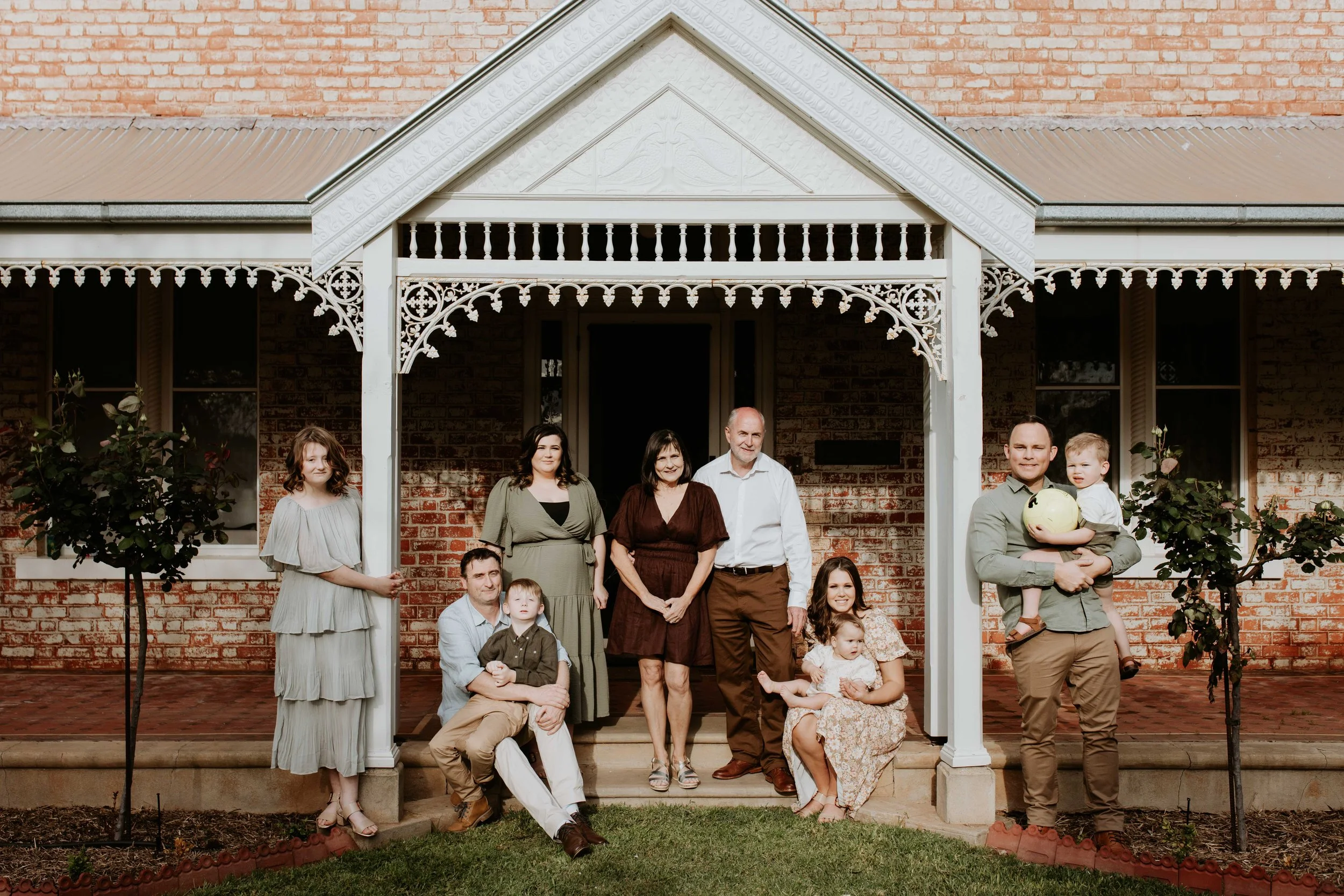 Family photo of ten people on a porch with painted brick wall and two small trees, some seated and others standing, some holding children, outdoors in daytime.