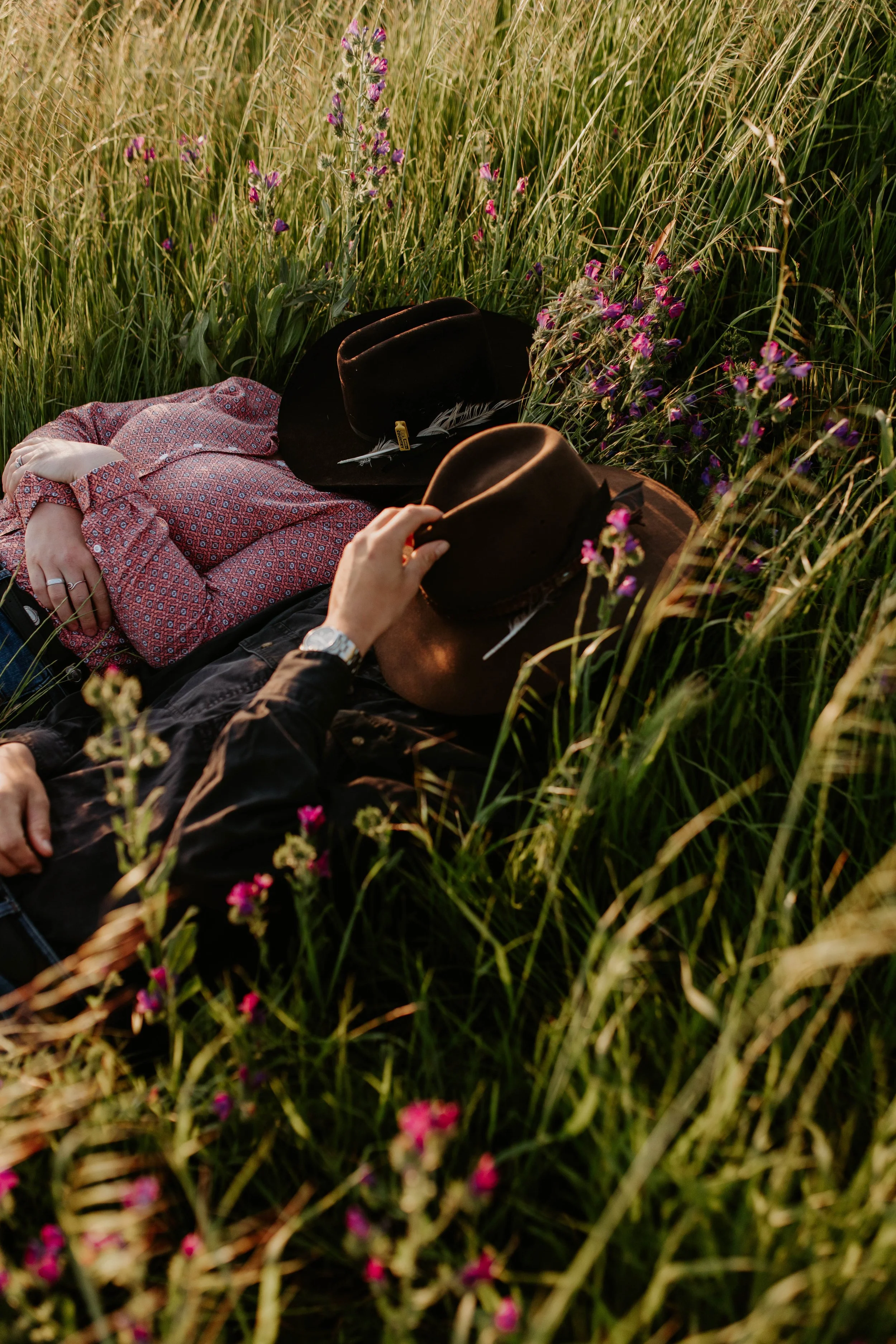 Two people lying in tall grass with wildflowers, wearing hats and casual clothing, enjoying a peaceful moment in nature.