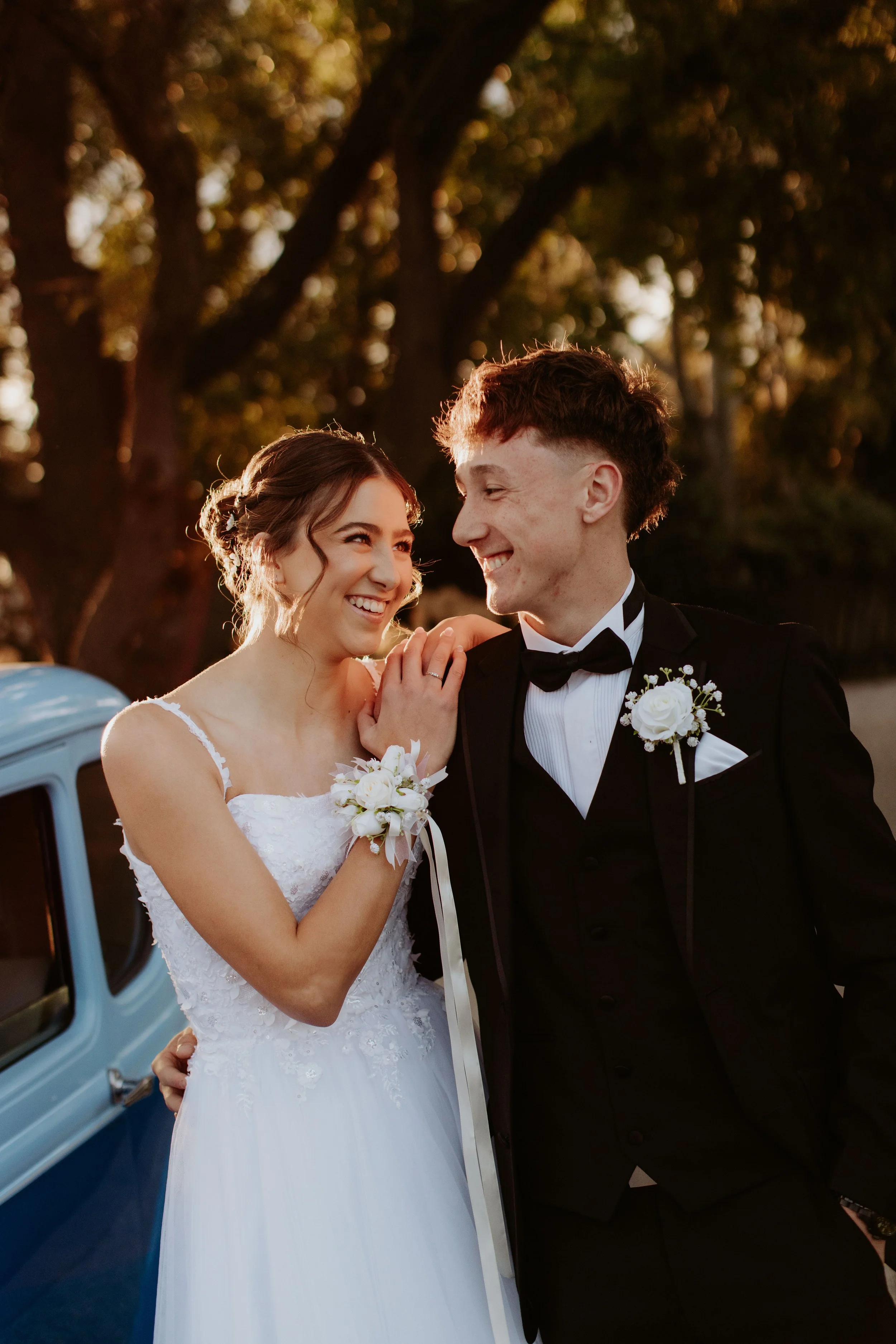 Deb couple smiling and looking at each other, outdoors during sunset, with a vintage car in the background. Modern Country Estate, Shepparton.