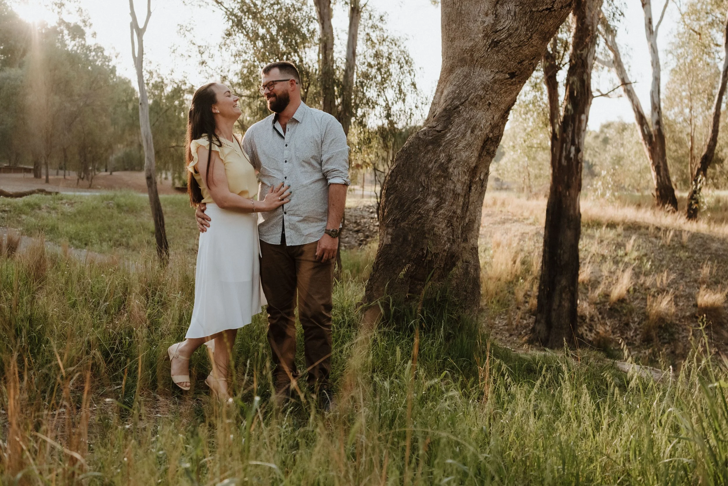 A couple standing affectionately in a grassy field near trees during sunset.
