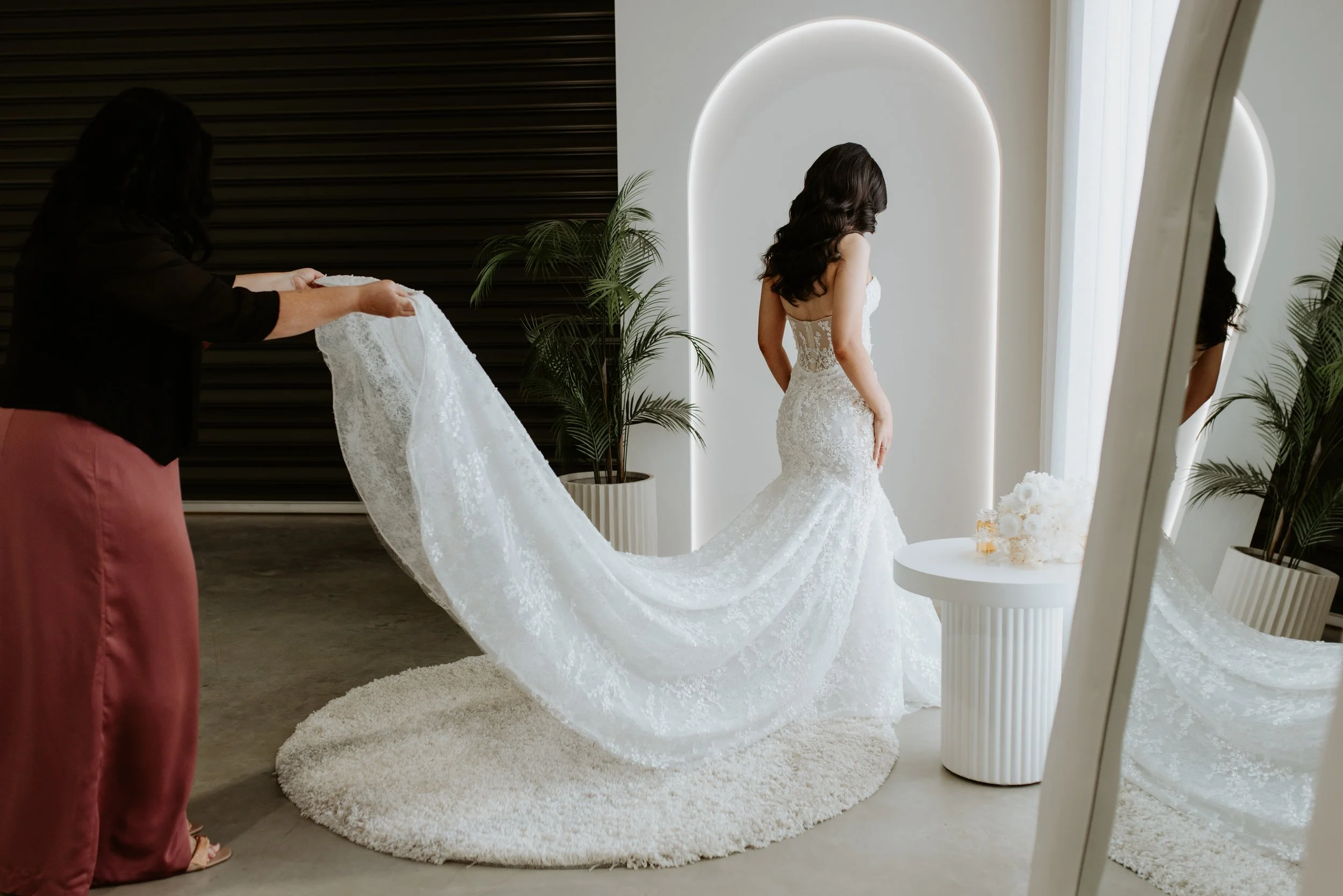 A woman in a wedding dress stands near a mirror while another woman helps with the train in a bright, modern room with plants and minimalist decor.