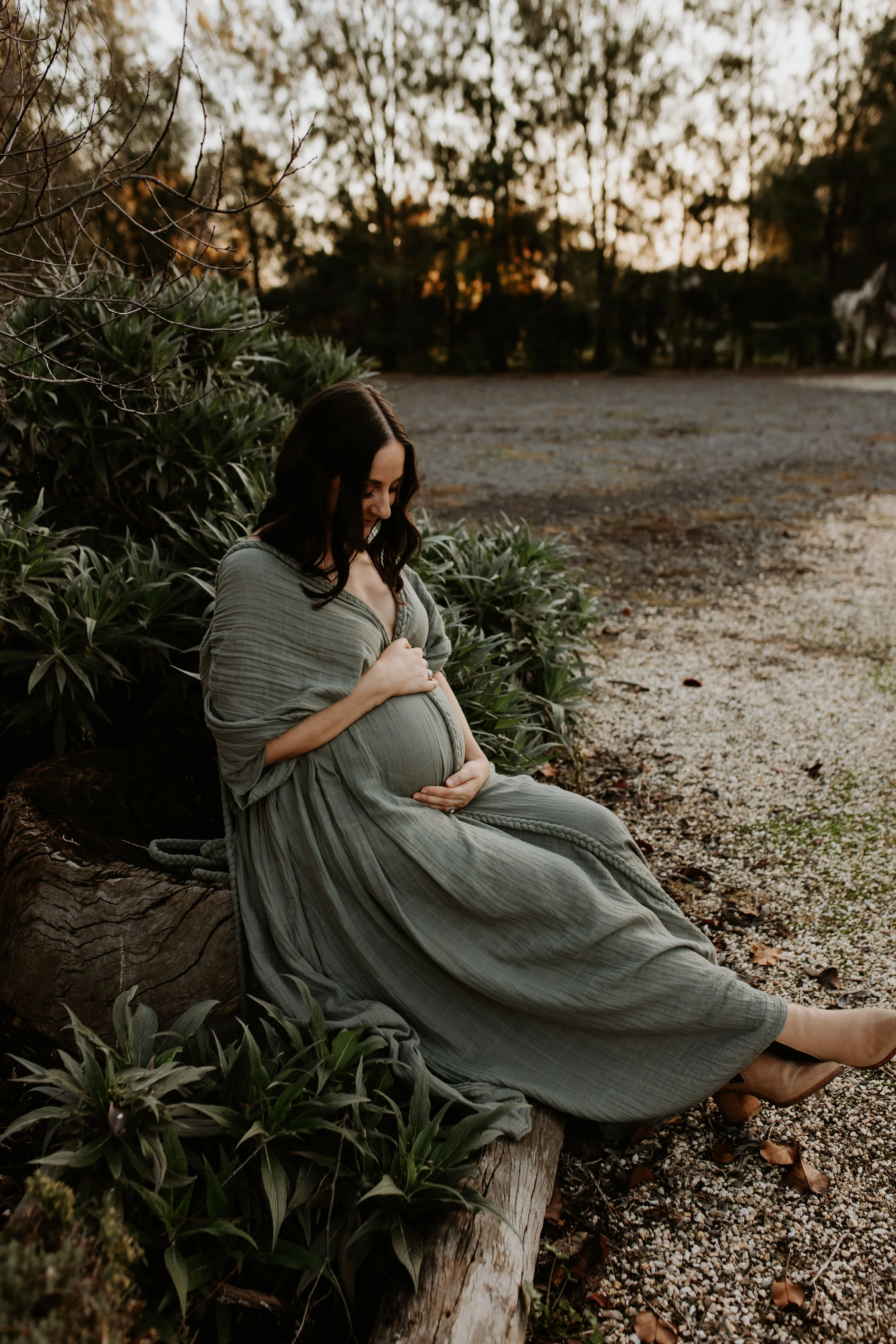 Pregnant woman sitting on a large log outdoors at sunset, cradling her belly and smiling gently.