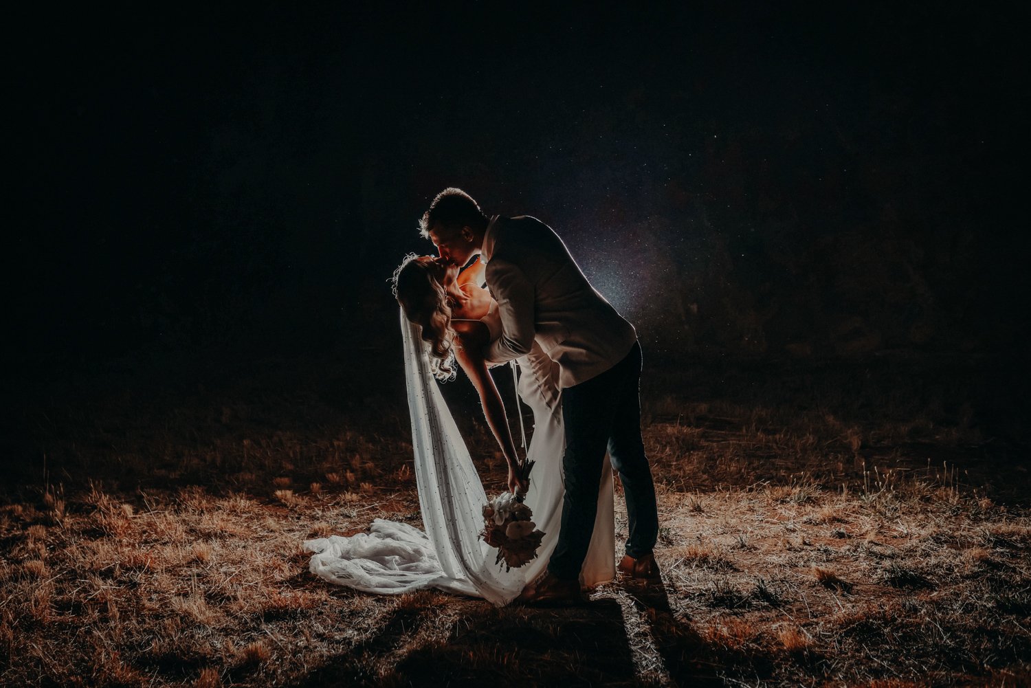 A bride and groom sharing a kiss at night, with the groom lifting the bride in a romantic pose, illuminated by backlighting against a dark background.