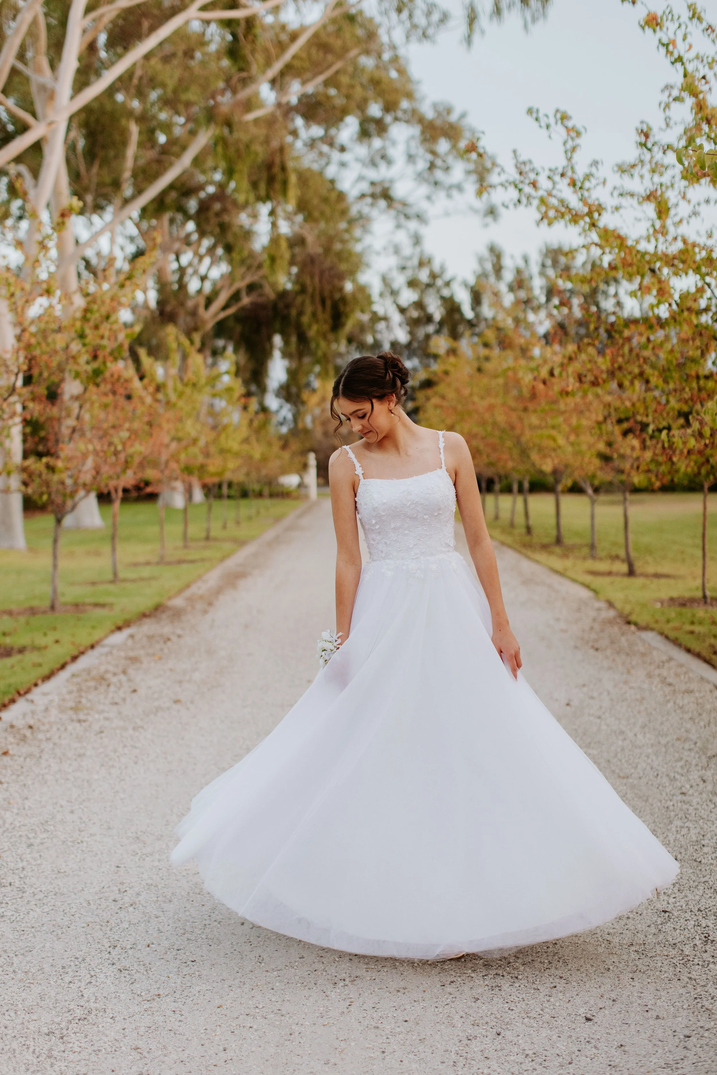 Young woman in a white dress standing on a gravel path lined with trees with autumn foliage. Modern Country Estate, Shepparton.