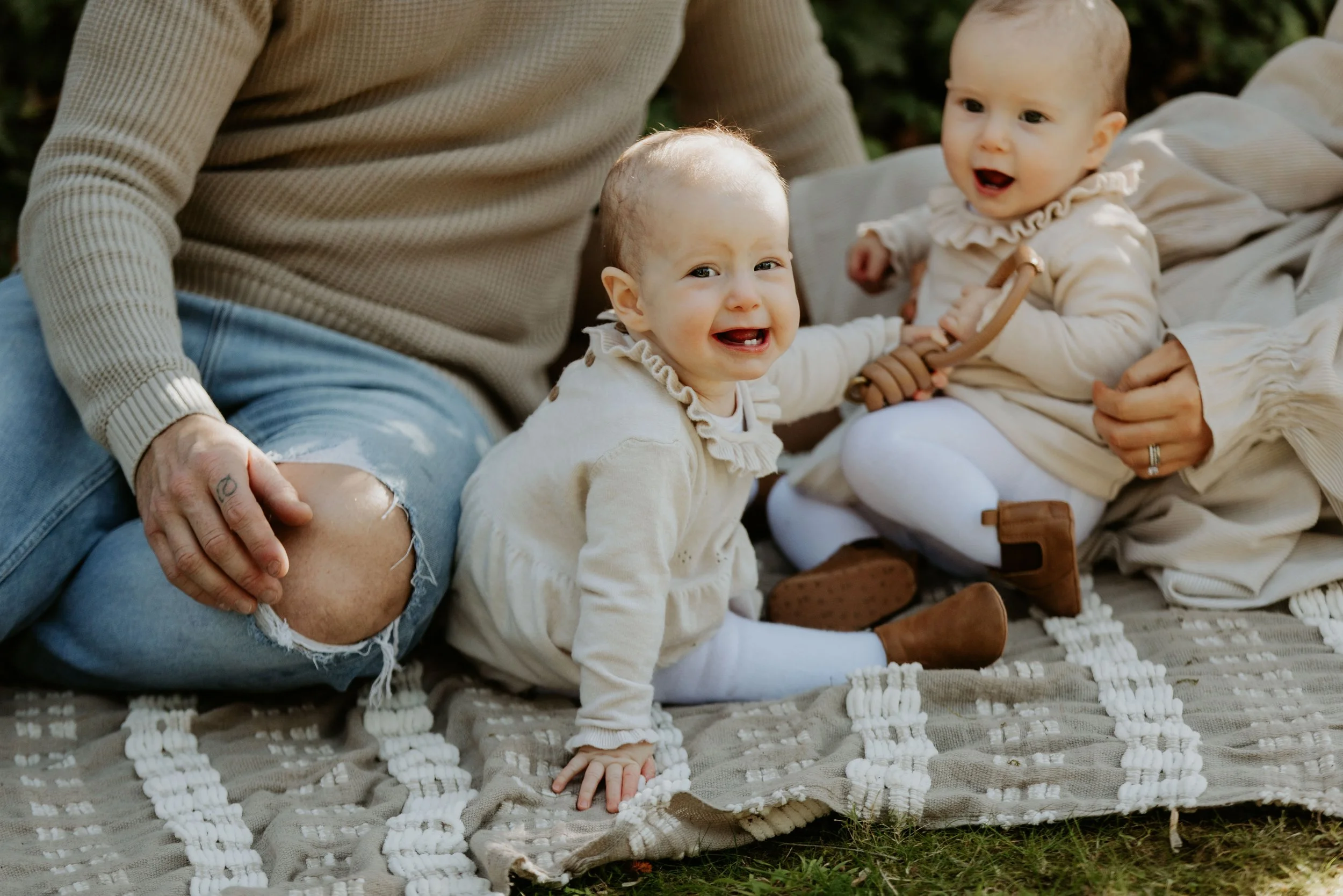 Two babies with light-colored clothing sitting on a blanket outdoors, smiling, with an adult's hand visible. One baby is reaching towards the adult, and the other is holding a small wooden toy.