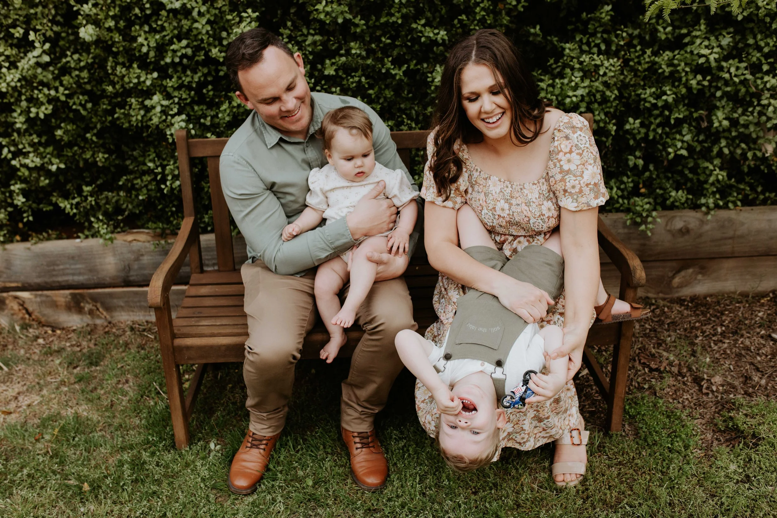 A family of four sitting on a wooden bench outdoors, enjoying a moment together. The father holds a young girl, the mother is playing with a boy who is hanging upside down, and they are all smiling and laughing.