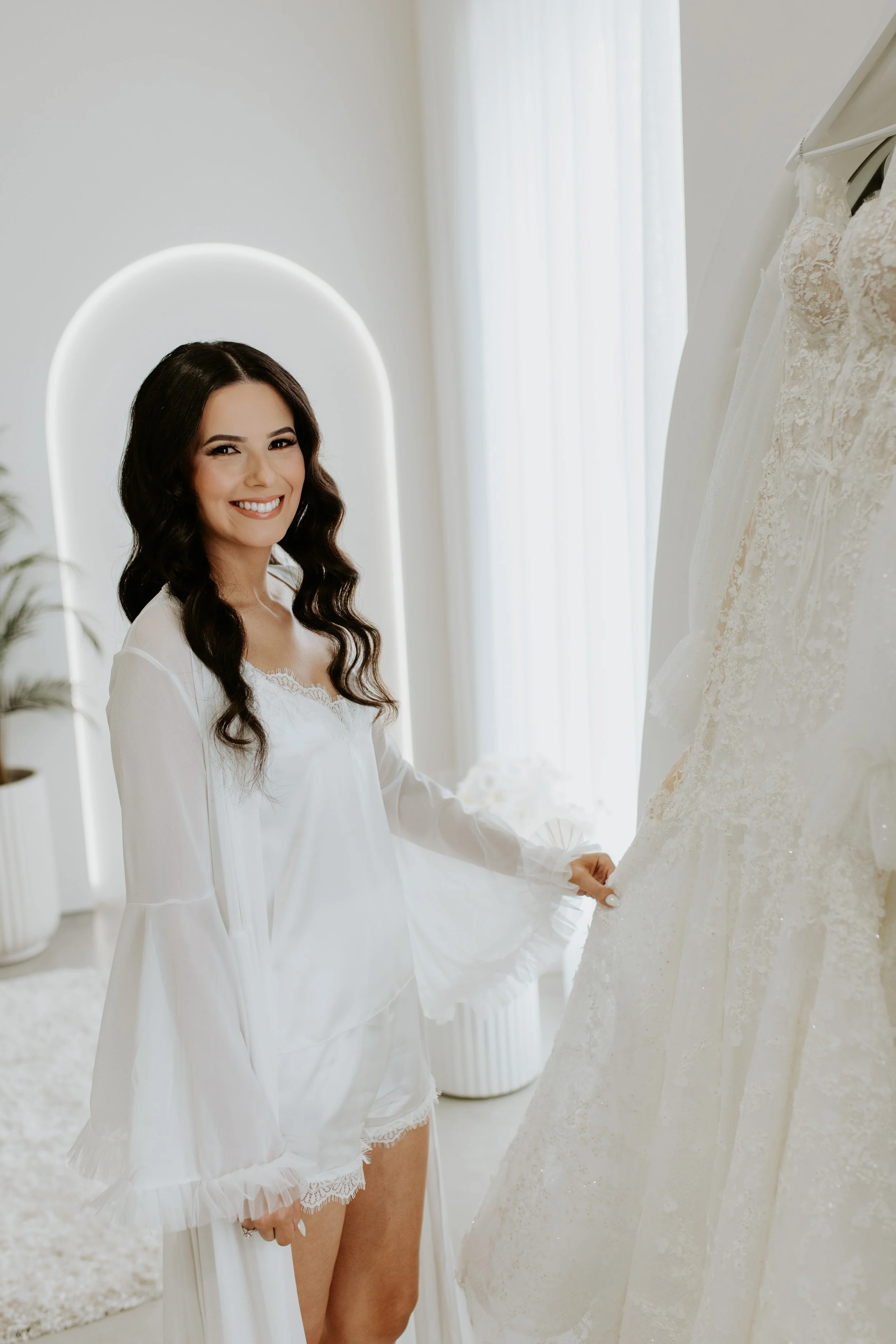 A woman with long dark wavy hair smiling while standing next to a wedding dress on a rack in a bright, modern room with white decor.