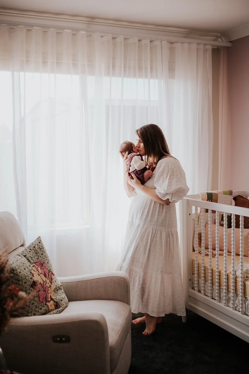 A woman in a white dress holding a newborn baby near a bright window with sheer curtains in a cozy room with a couch and a crib.