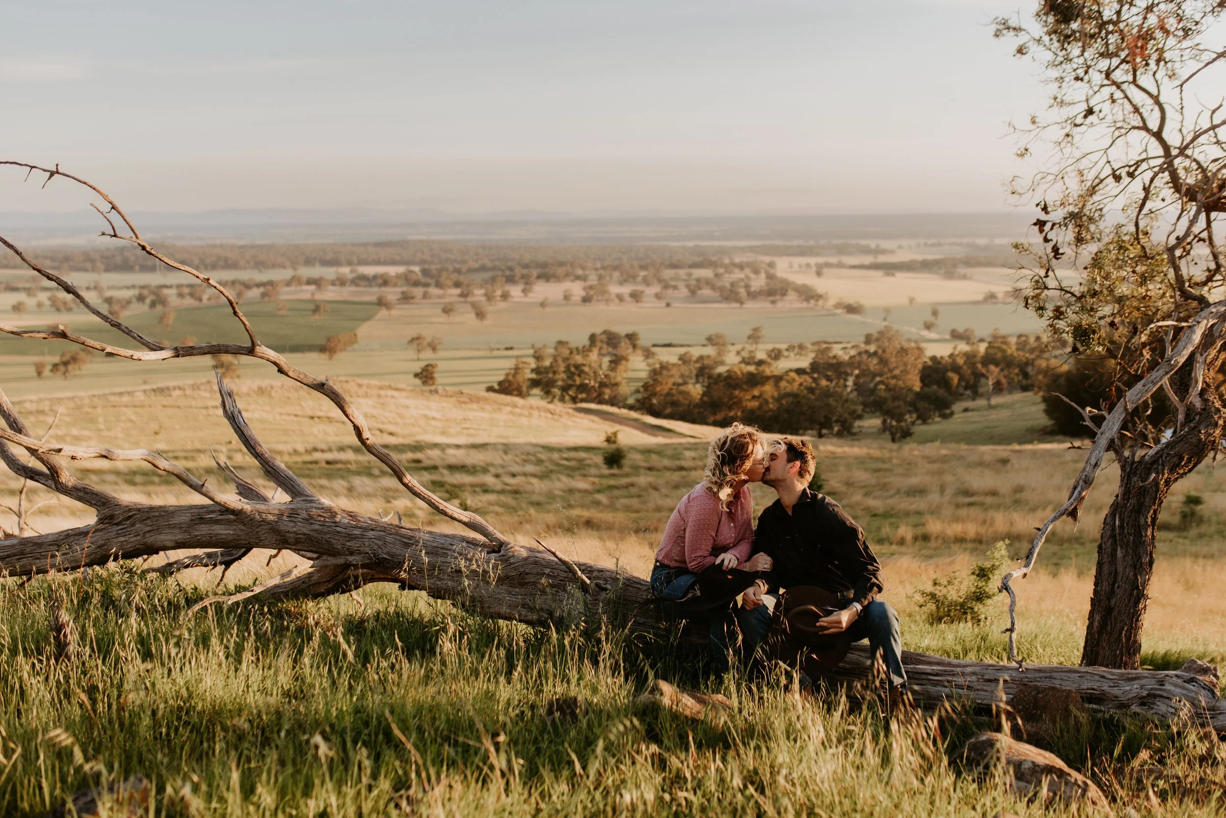 A couple sitting on a fallen tree, kissing with a scenic countryside landscape in the background.