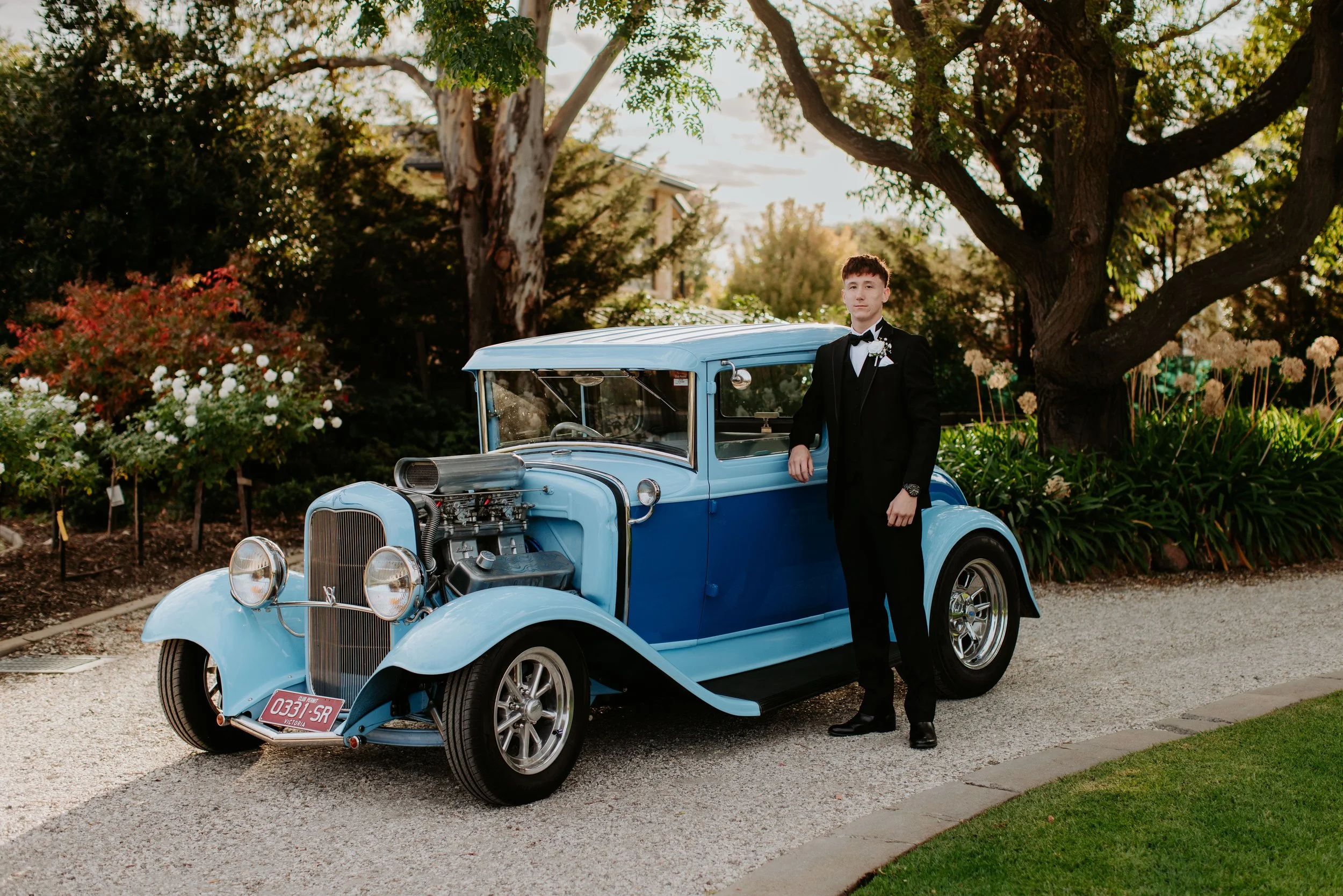 A young man in a black tuxedo standing next to a vintage blue car in a garden with trees and flowers.