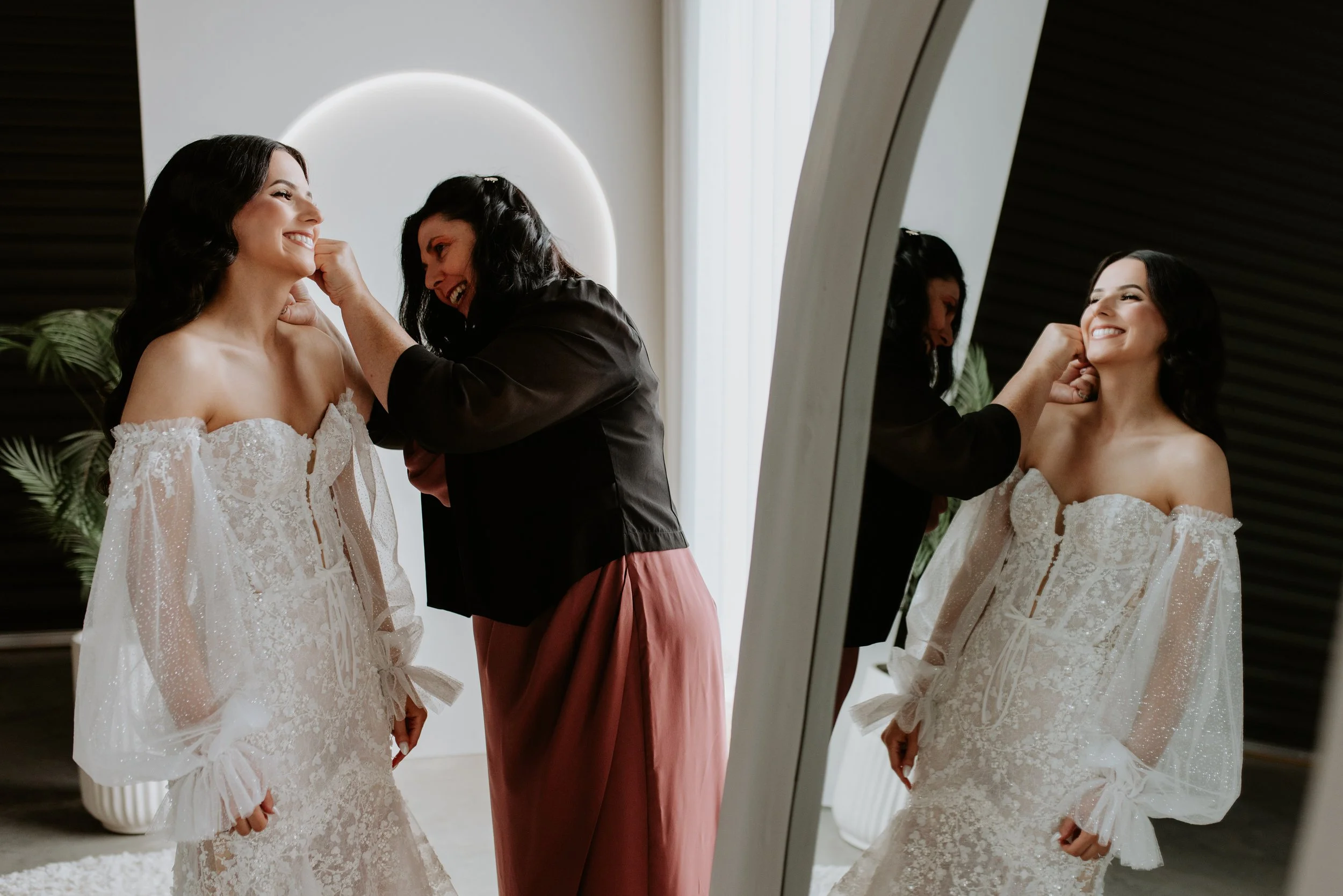 A woman in a wedding dress smiles as a makeup artist touches up her face, with her reflection visible in a mirror.