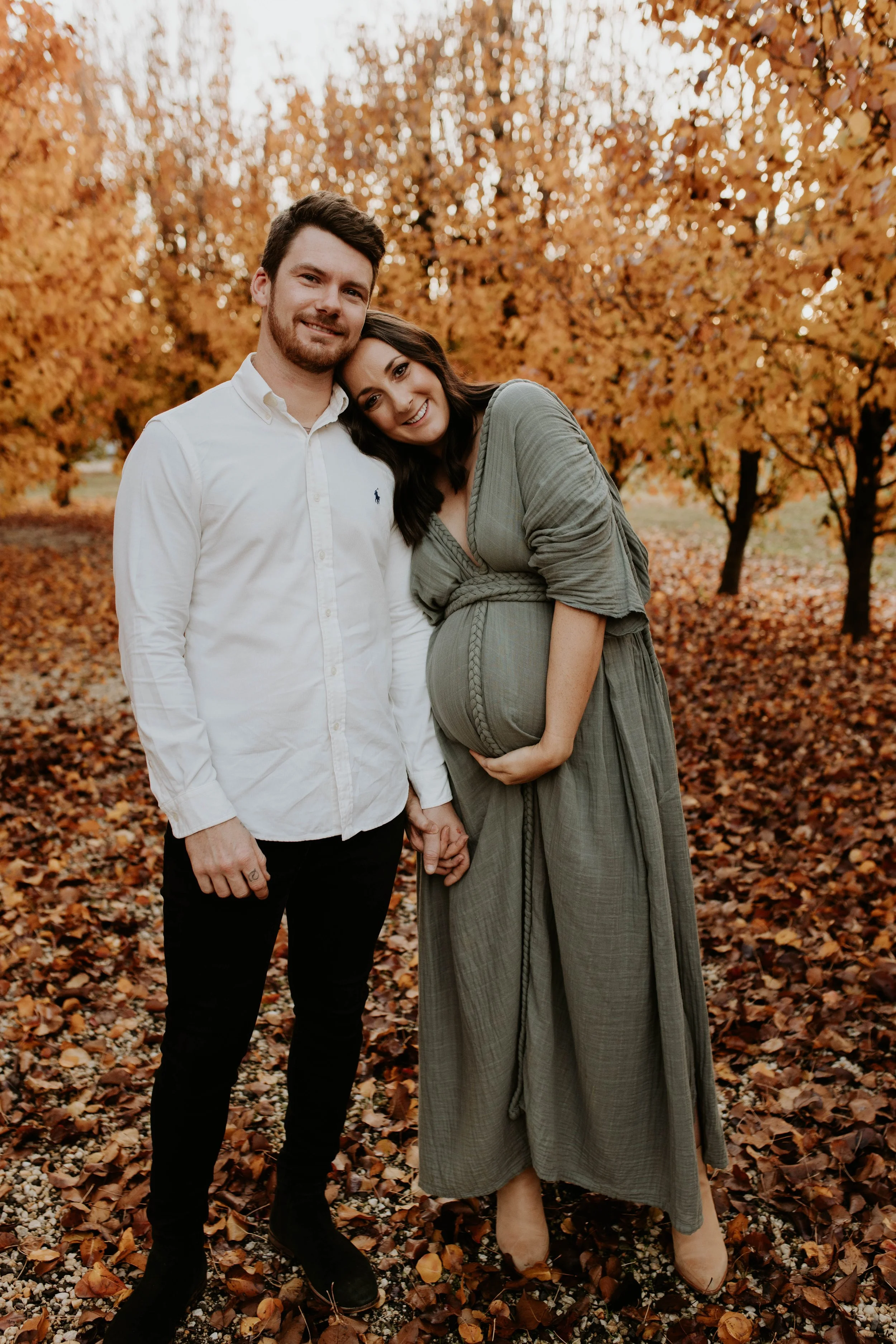 A happy couple holding hands outdoors with autumn leaf-covered ground and orange trees in the background. The pregnant woman is wearing a long green dress and the man is dressed in a white shirt and black pants.