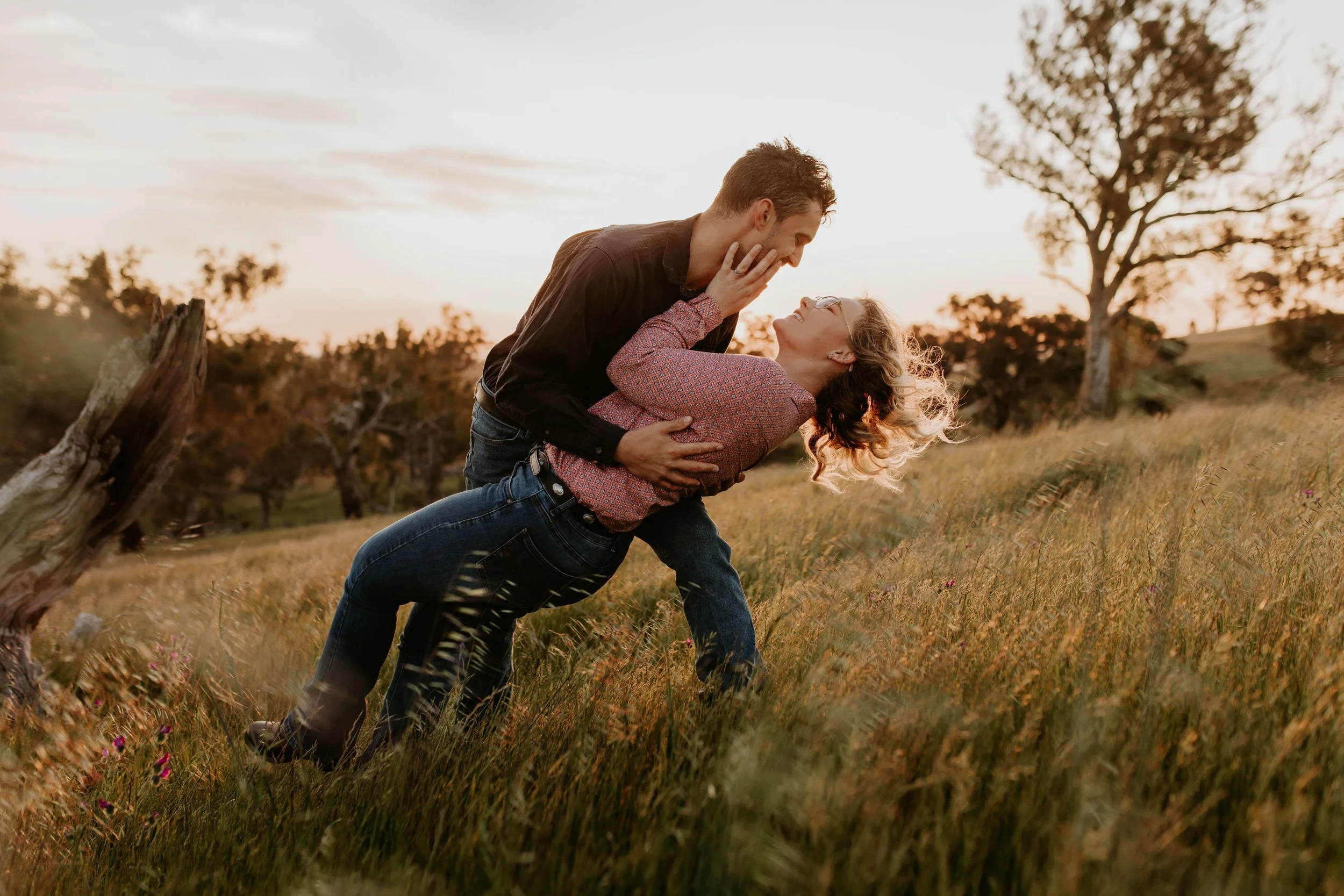 A couple is dancing and embracing in a grassy field during sunset, with trees in the background.
