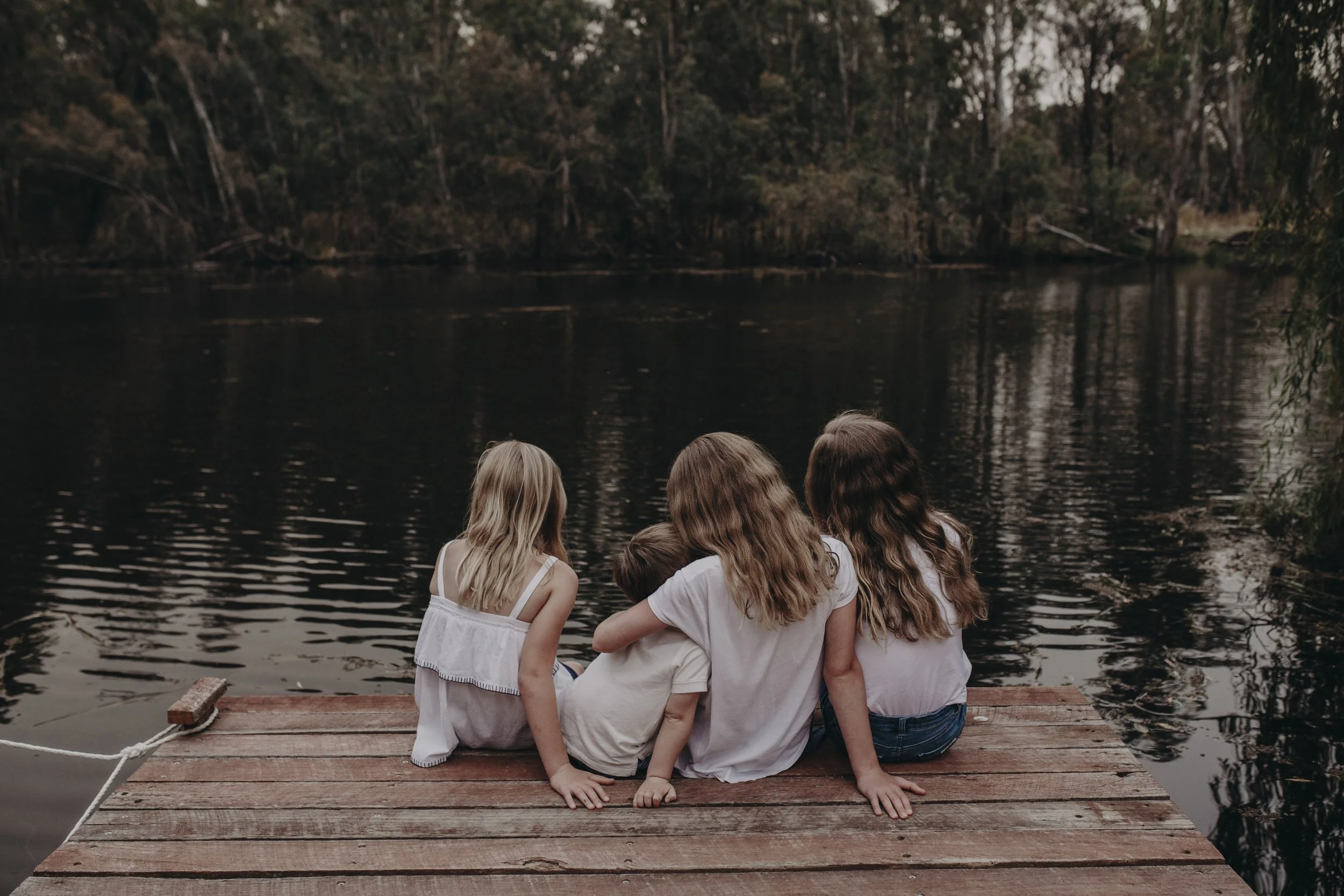 Four children sitting on a wooden dock by a lake, facing away from the camera, surrounded by trees.