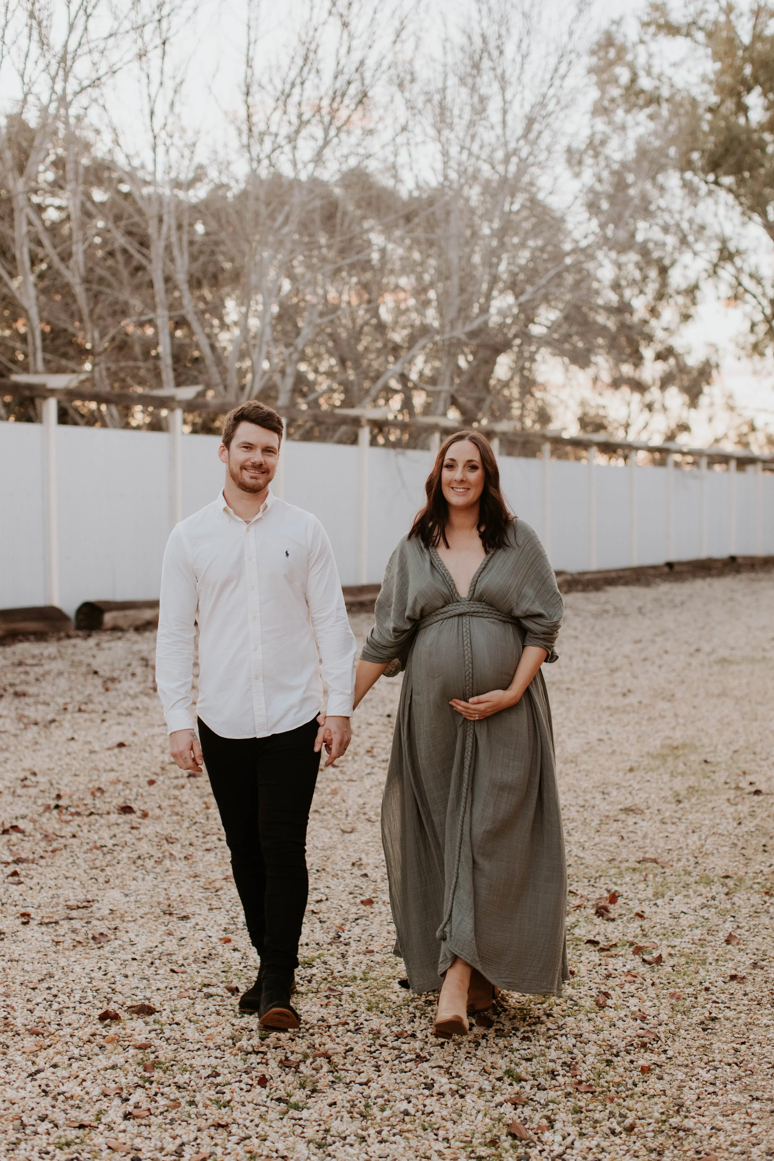 A pregnant woman in a long gray dress holding her belly walking outdoors with a man in a white shirt and black pants, smiling together on a gravel path background with trees and a white fence.