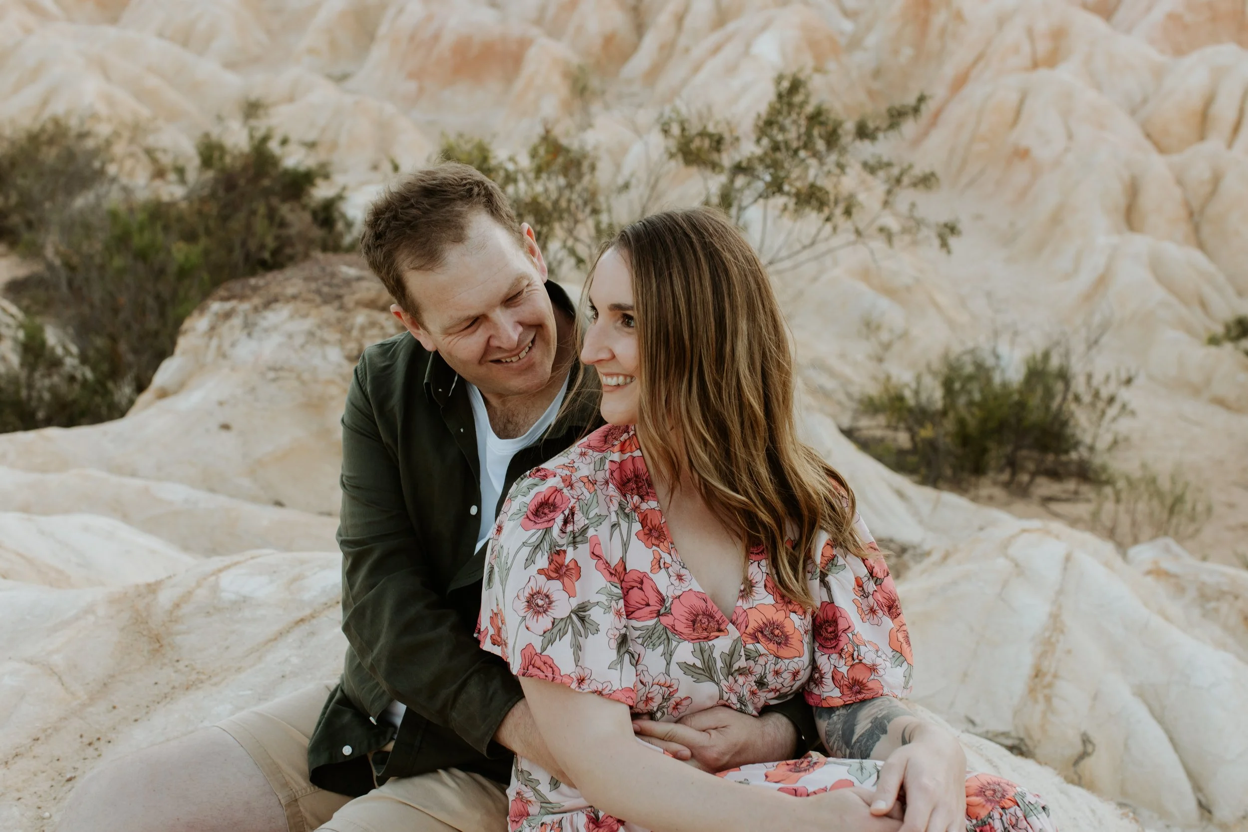 A happy couple sitting on rocks in a desert landscape, smiling at each other.