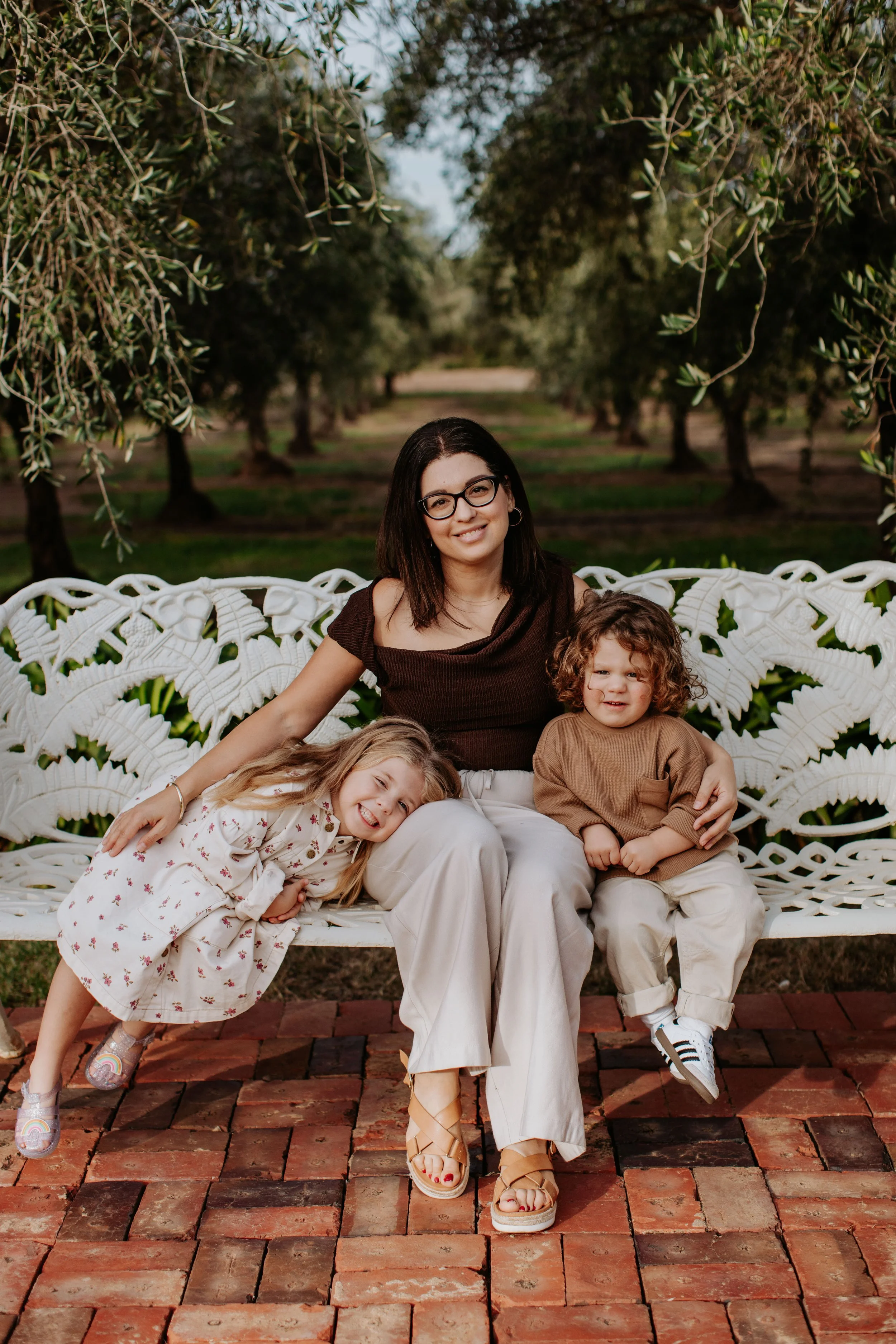 A woman with dark hair and glasses sitting on a decorative white garden bench outdoors, with two young children, a girl with blonde hair in a floral dress leaning on her and a curly-haired boy in a brown shirt sitting beside her, all smiling.