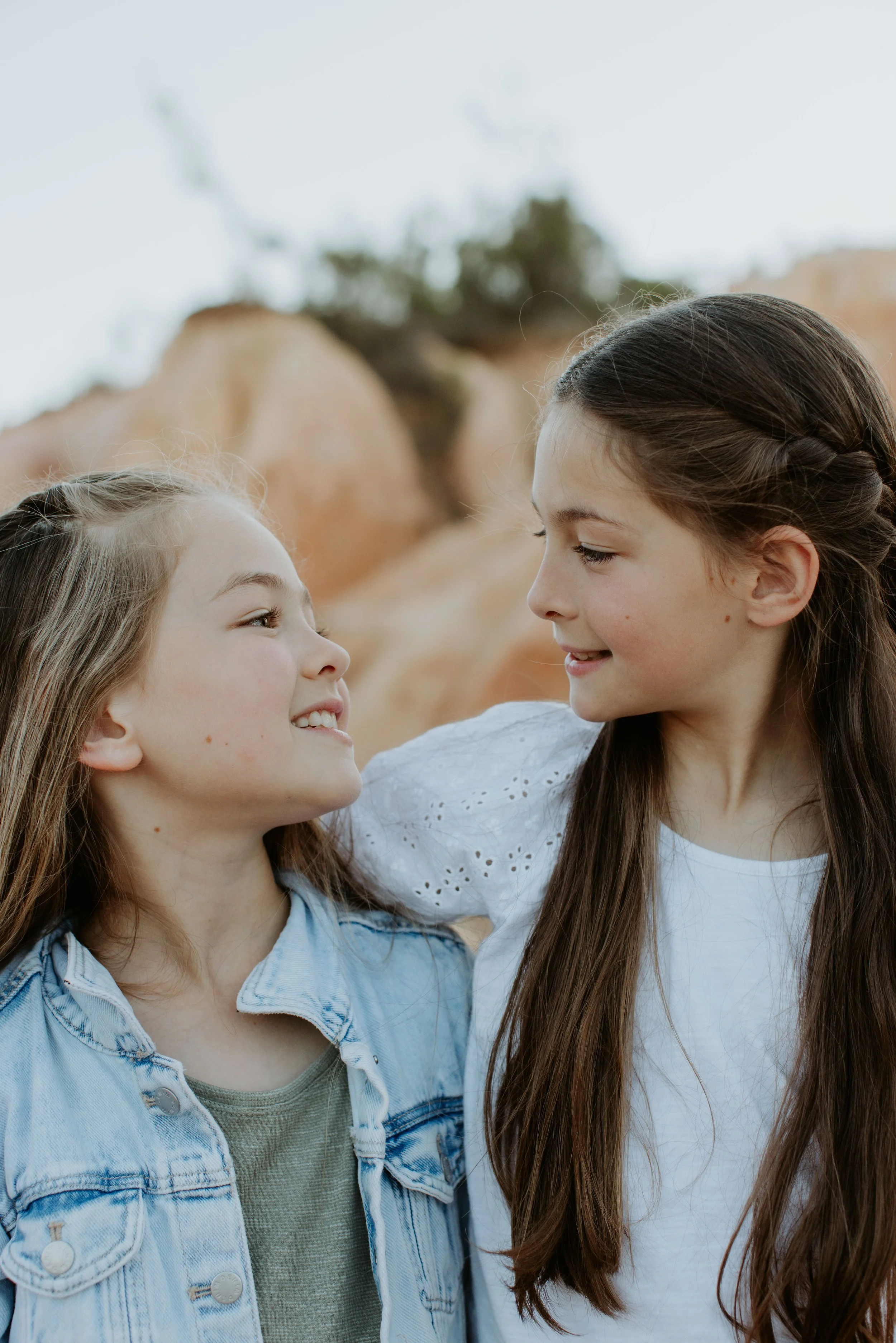 Two young girls smiling at each other outdoors near rocks with a cloudy sky.