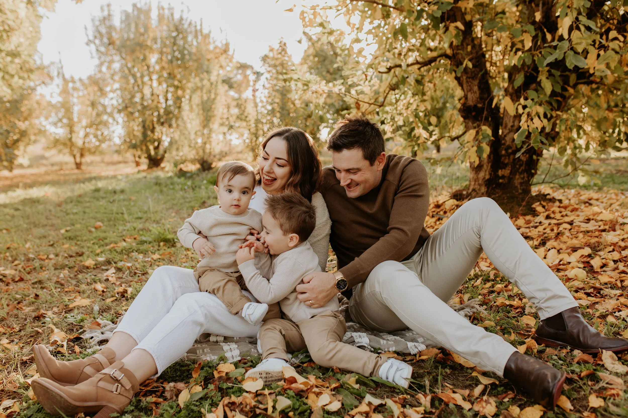 Family sitting on the ground in autumn park, smiling and playing with children near a large tree with fall leaves.