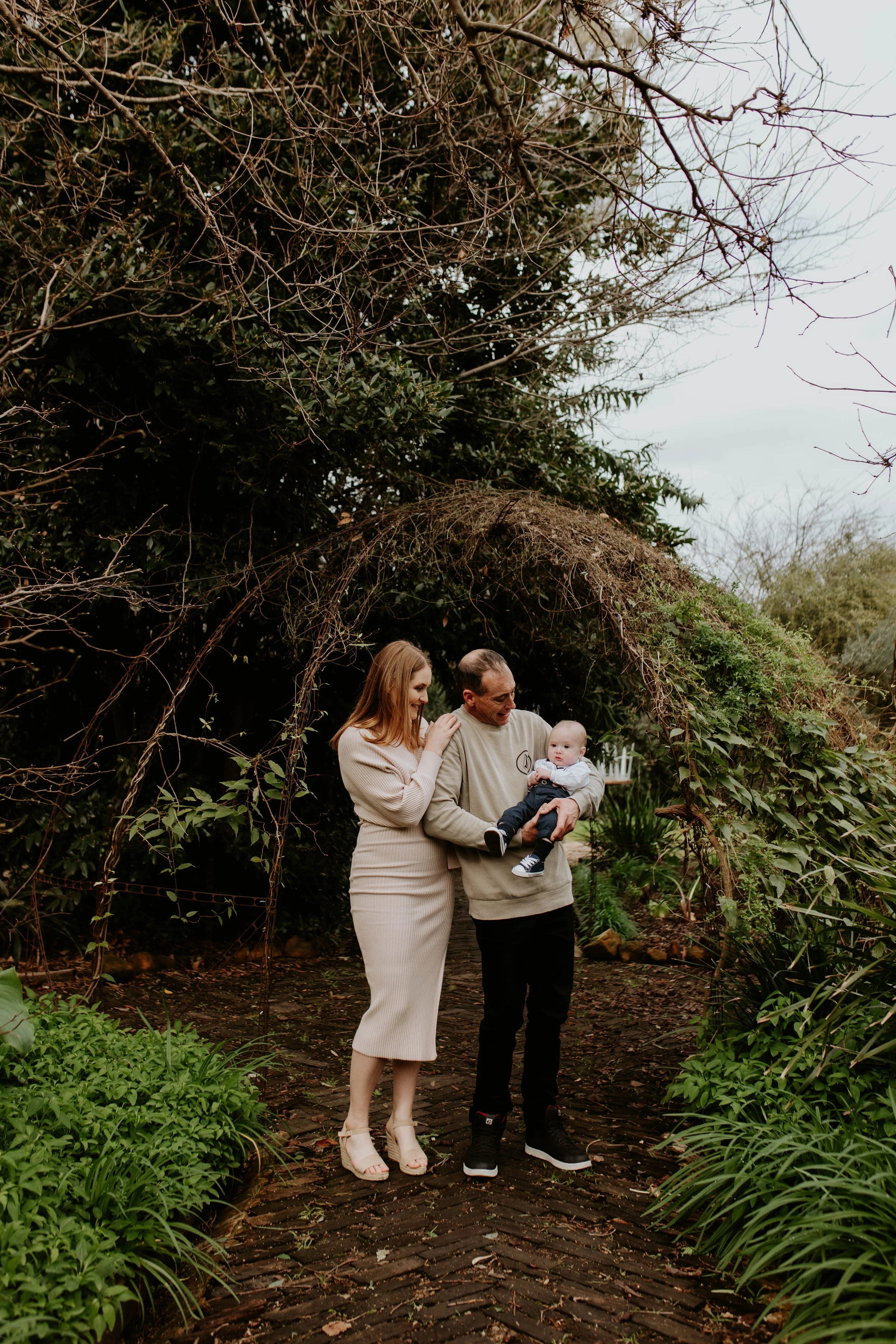 A family of three standing on a brick walkway in a lush garden with an archway overhead, the woman touching the man's shoulder and the man holding a baby boy wearing a white shirt and dark pants.