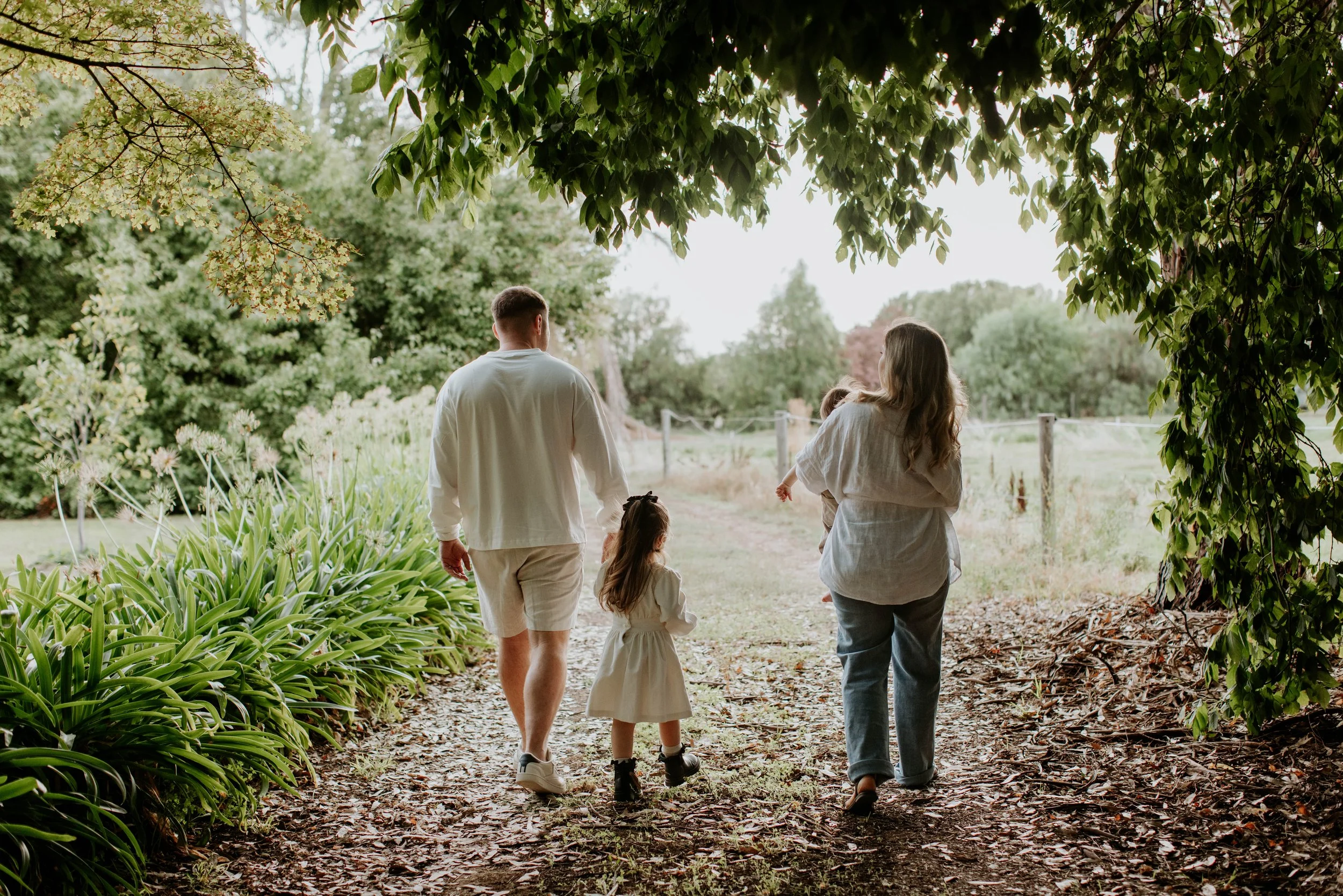 A family of four, including a man, woman, and two young children, walks along a garden path under the shade of trees in a lush, green outdoor setting.