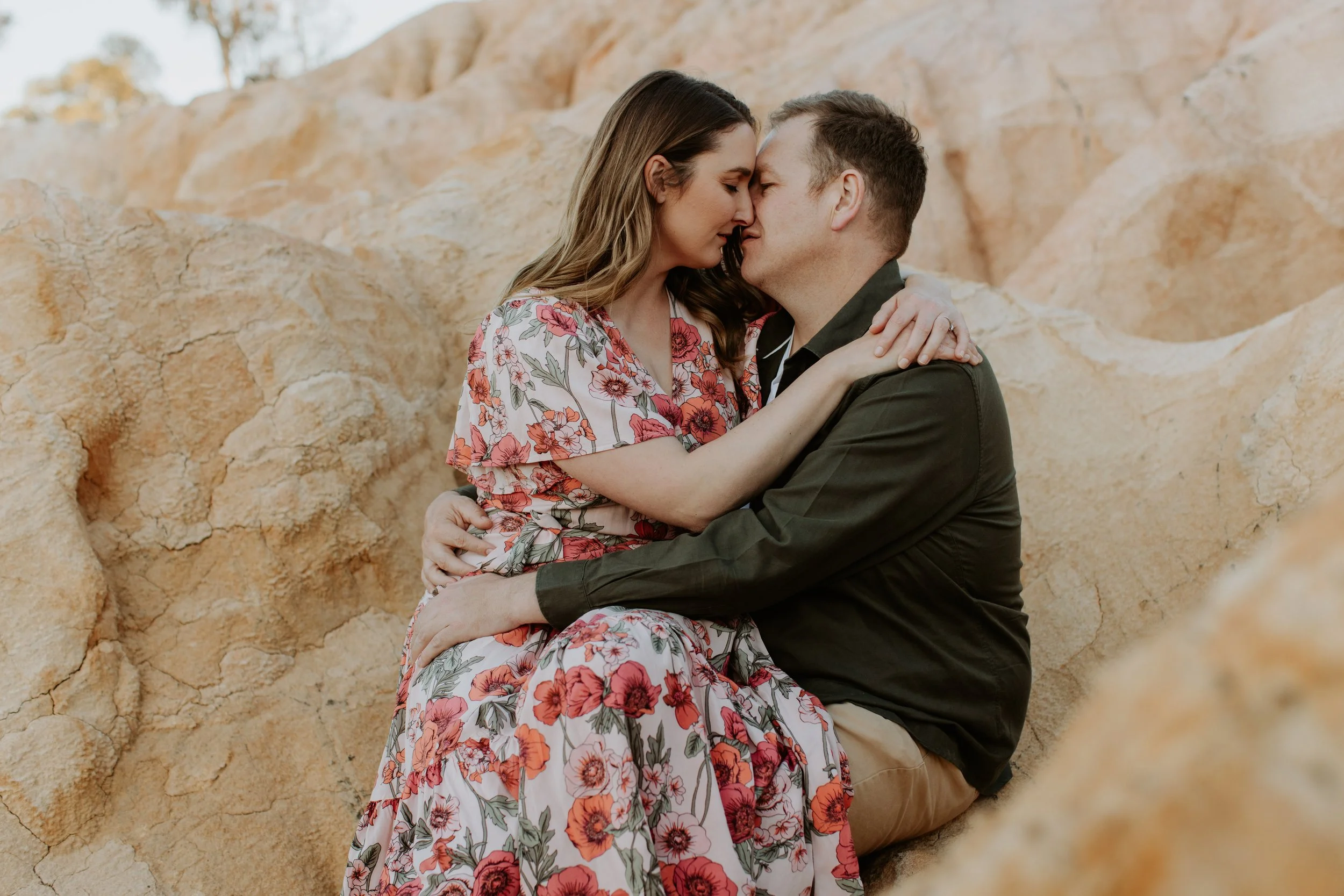 A couple sits close together on a rocky landscape, embracing and touching foreheads with eyes closed.