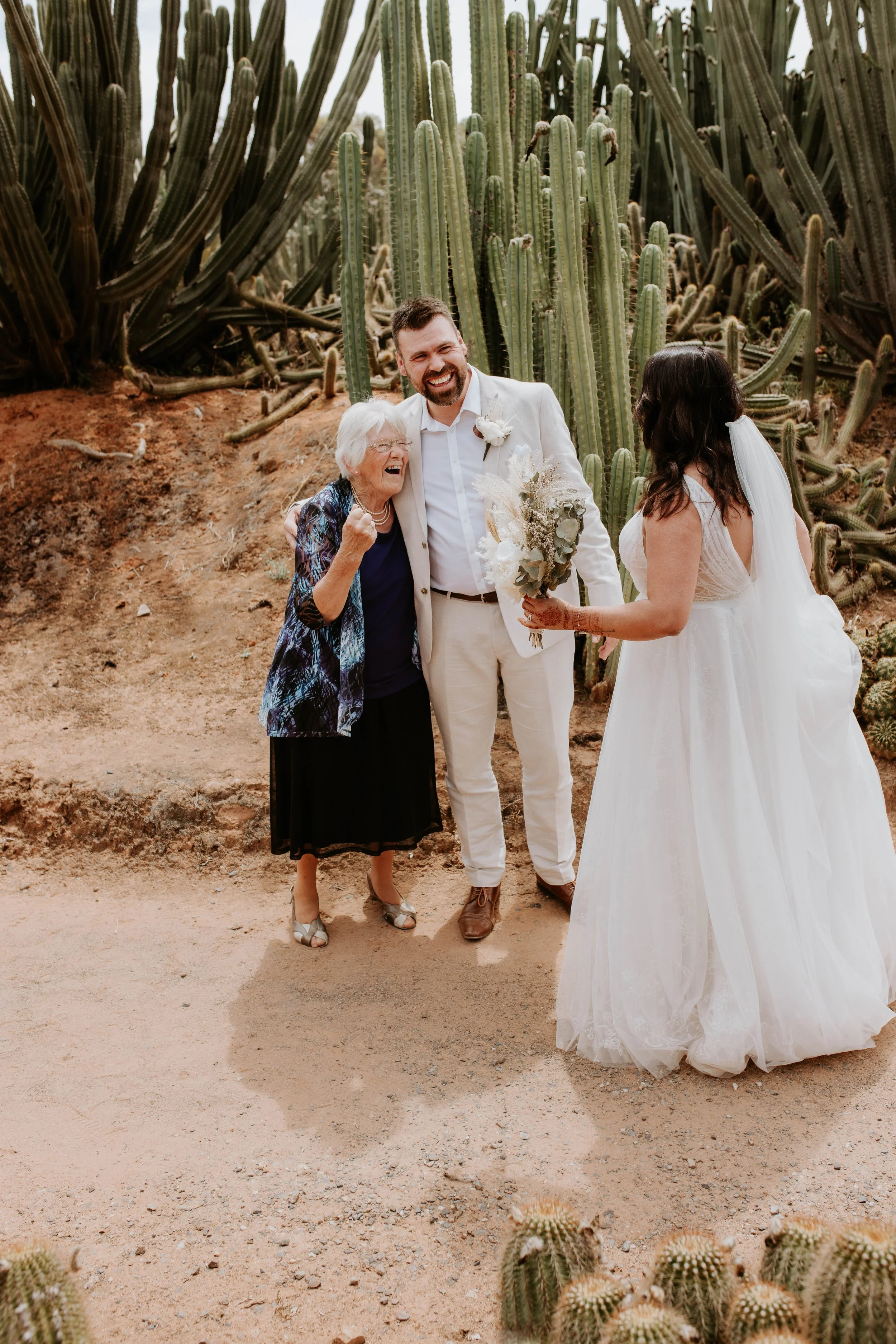 A bride, groom, and an older woman sharing a joyful moment outdoors in a desert setting with tall cacti in the background. Cactus Country.