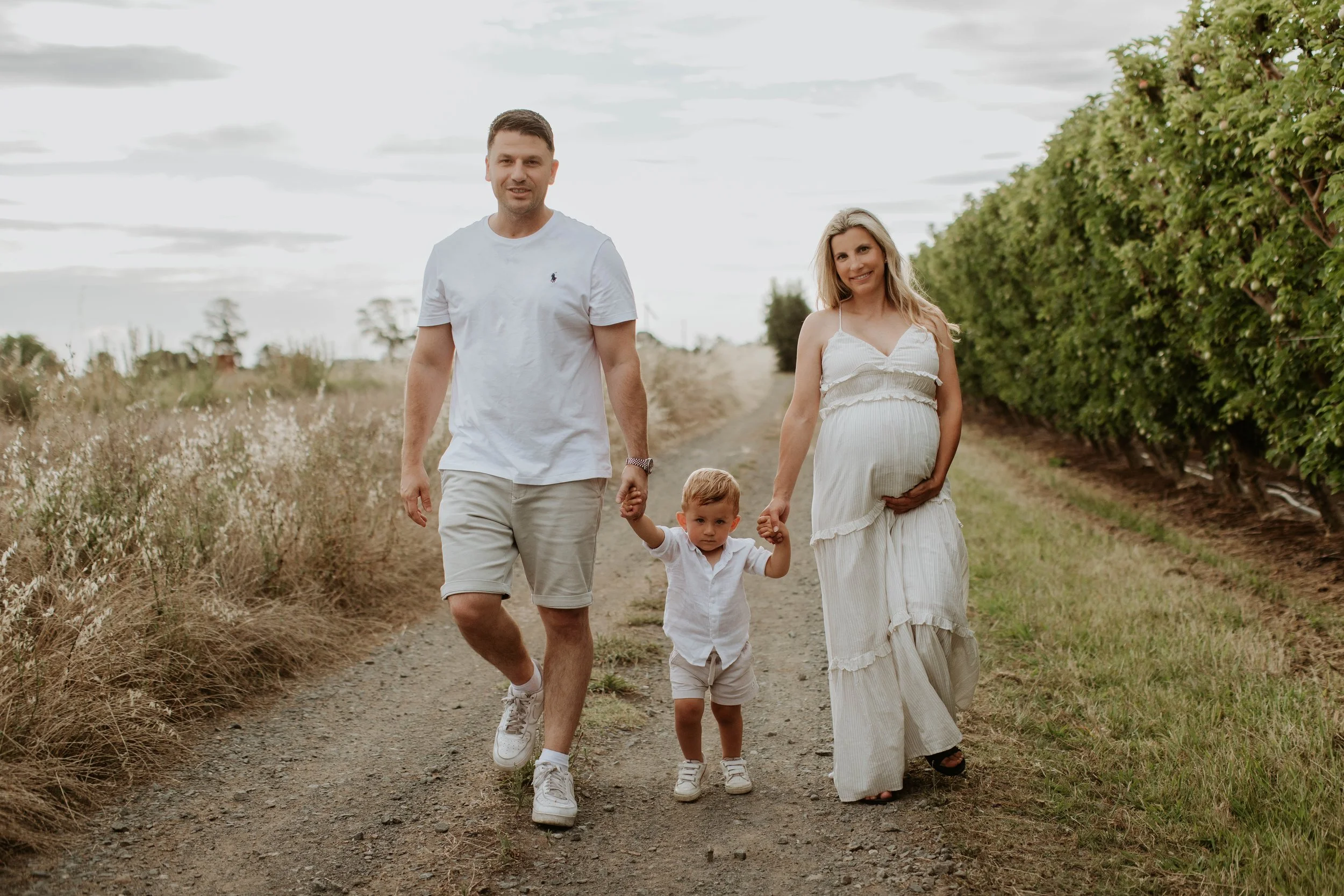A family of three walking down a rural dirt path, holding hands, with a pregnant mother on the right and a man and young boy on the left, surrounded by grass and green foliage.