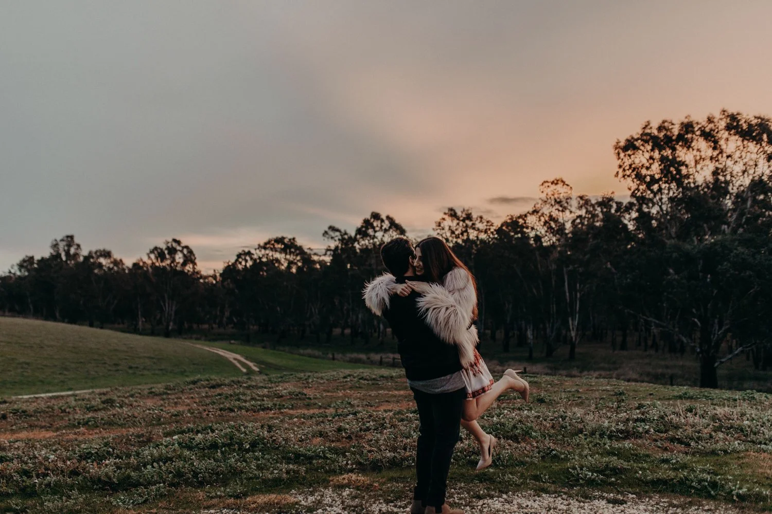 A couple happily embracing outdoors during sunset, with the woman being lifted by the man, surrounded by open grassy fields and a line of trees in the background.