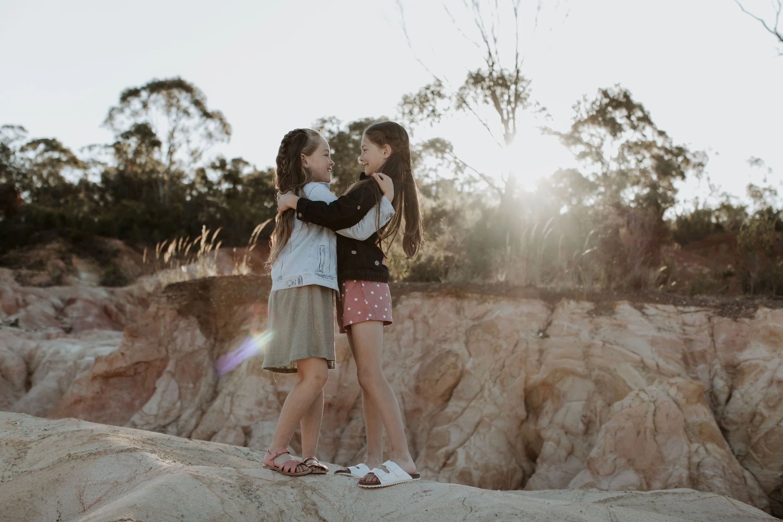 Two young girls hugging on a rocky outdoor landscape during sunset.
