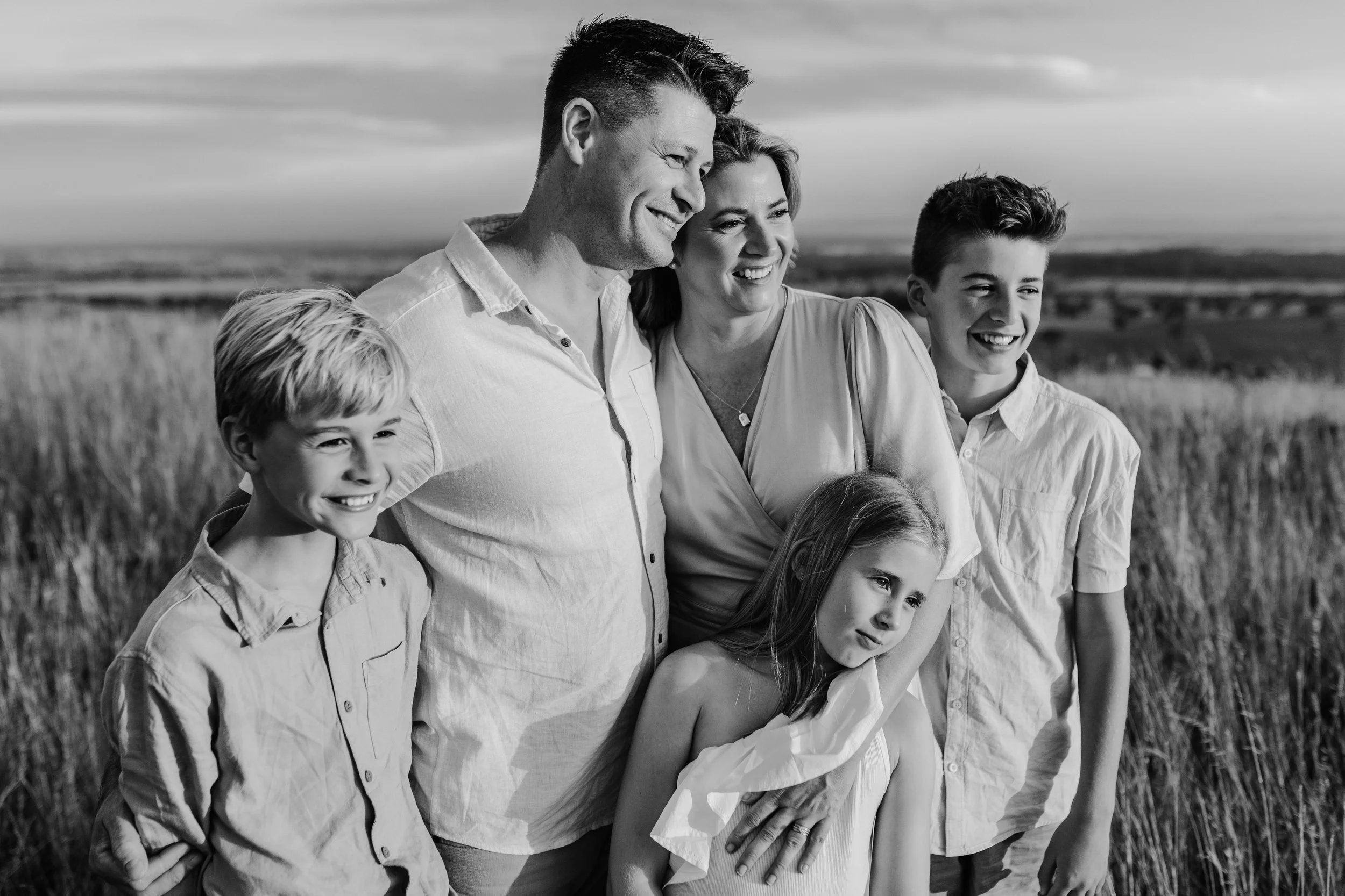 A family of six stands together in a field of tall grass outdoors, smiling and enjoying the moment. The group includes an adult man and woman, two teenage boys, a young boy, and a girl, all dressed in casual light-colored clothing.
