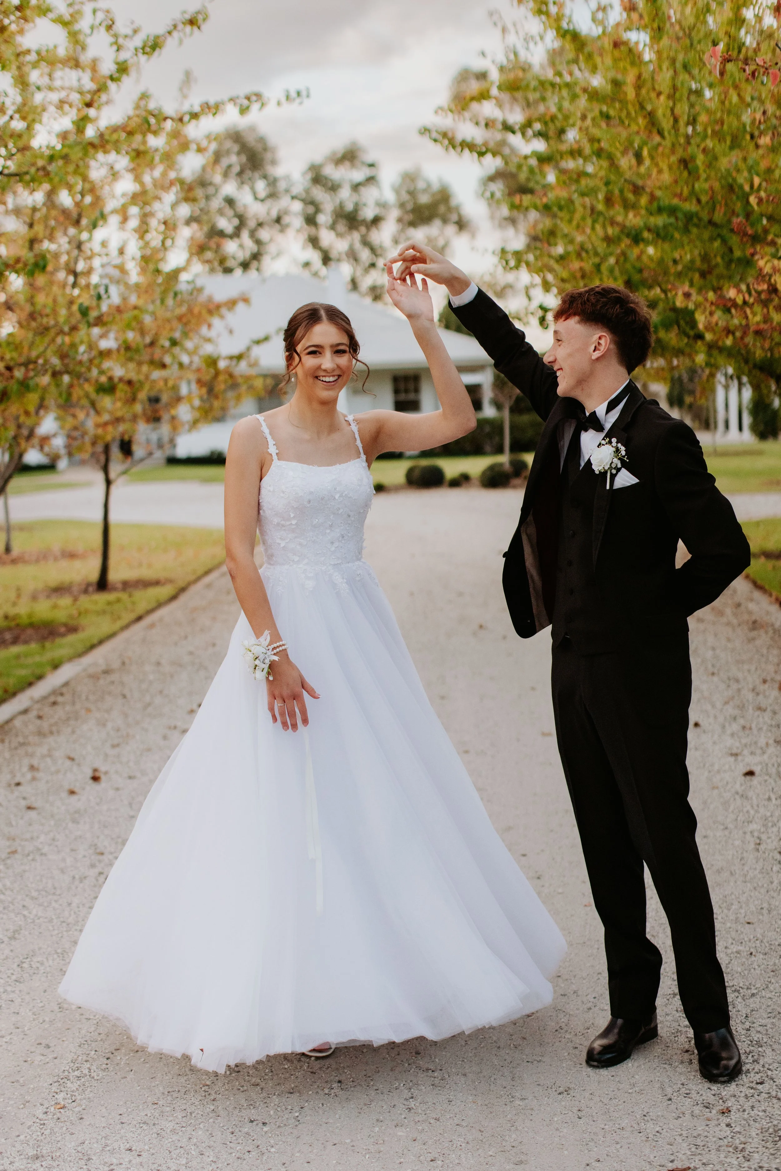 A deb couple dancing outdoors on a gravel pathway with trees and a white house in the background. Modern Country Estate, Shepparton.