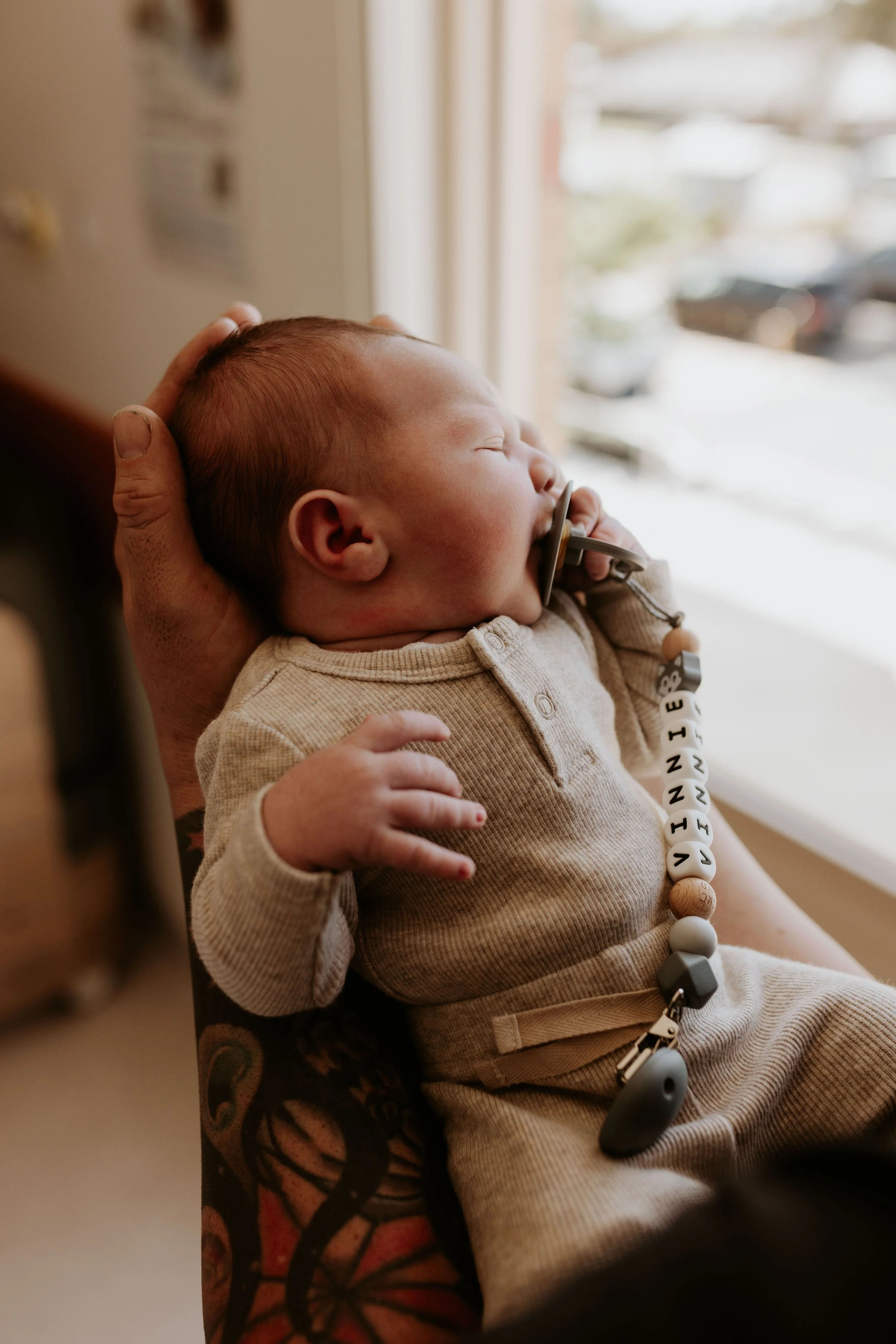 A baby with a pacifier, lying on an adult's arm, inside a room near a window, with cars visible outside.