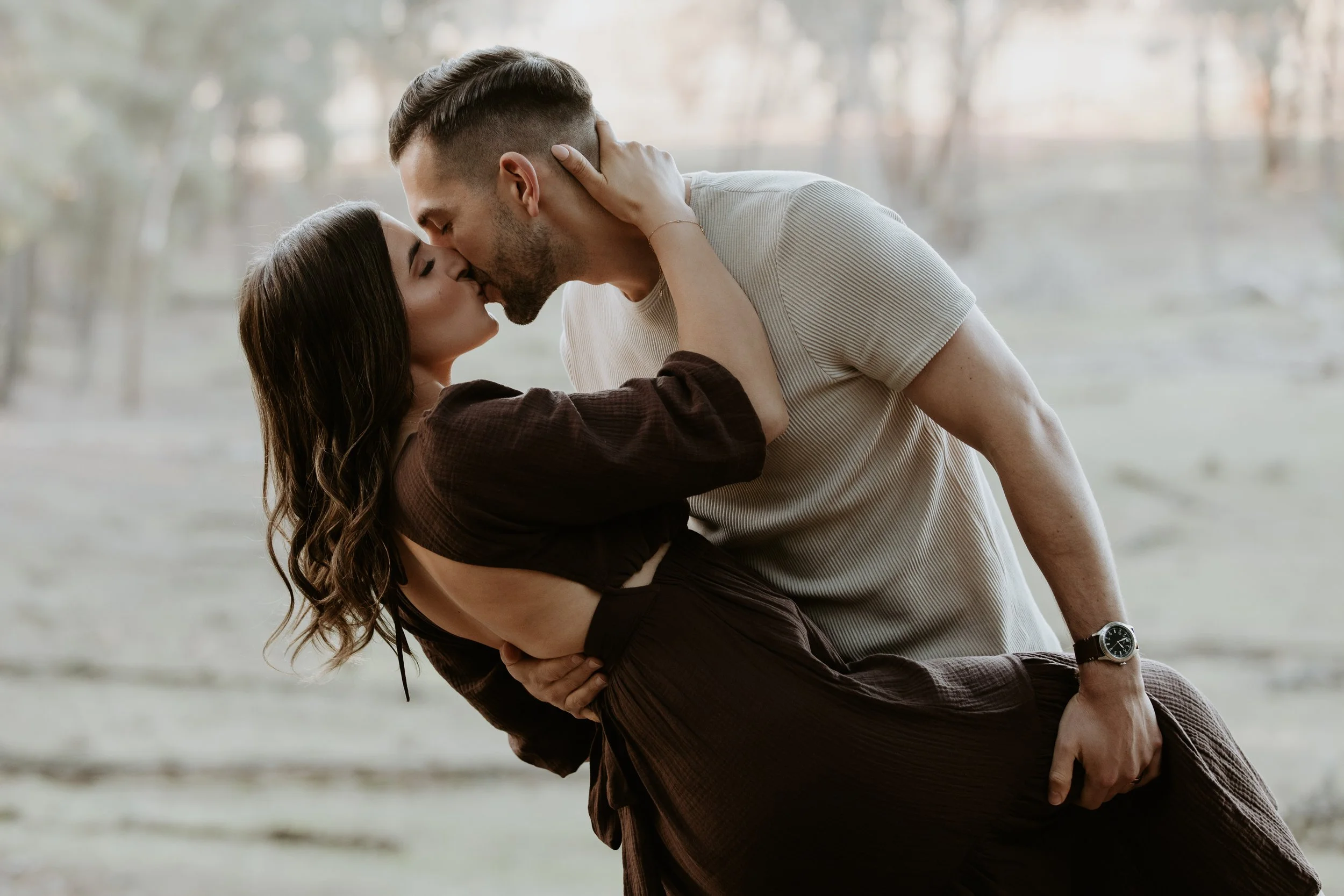 A couple sharing a romantic kiss outdoors during sunset, with the man holding the woman in his arms.
