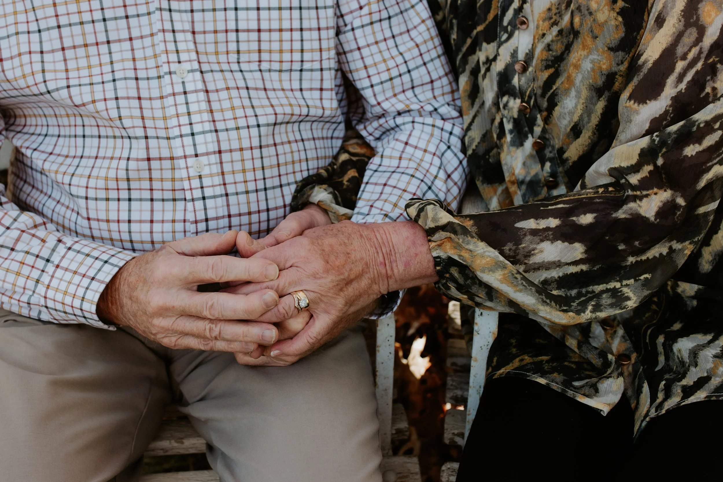 Close-up of two elderly people holding hands, one wearing a checkered shirt and the other wearing a patterned long-sleeve shirt, sitting outdoors.
