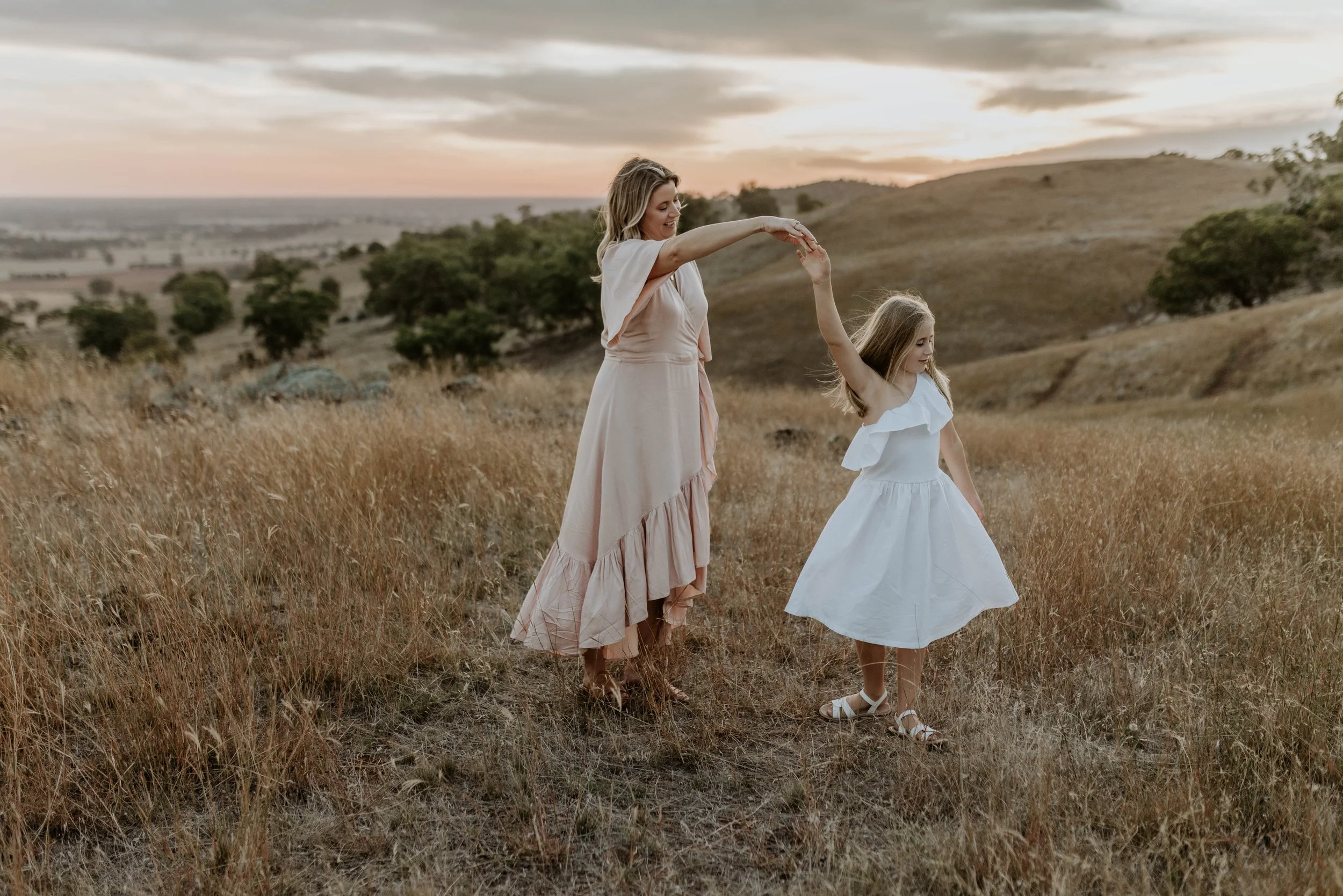 A woman and a young girl dancing in a grassy field at sunset, with a scenic landscape of rolling hills and trees in the background.
