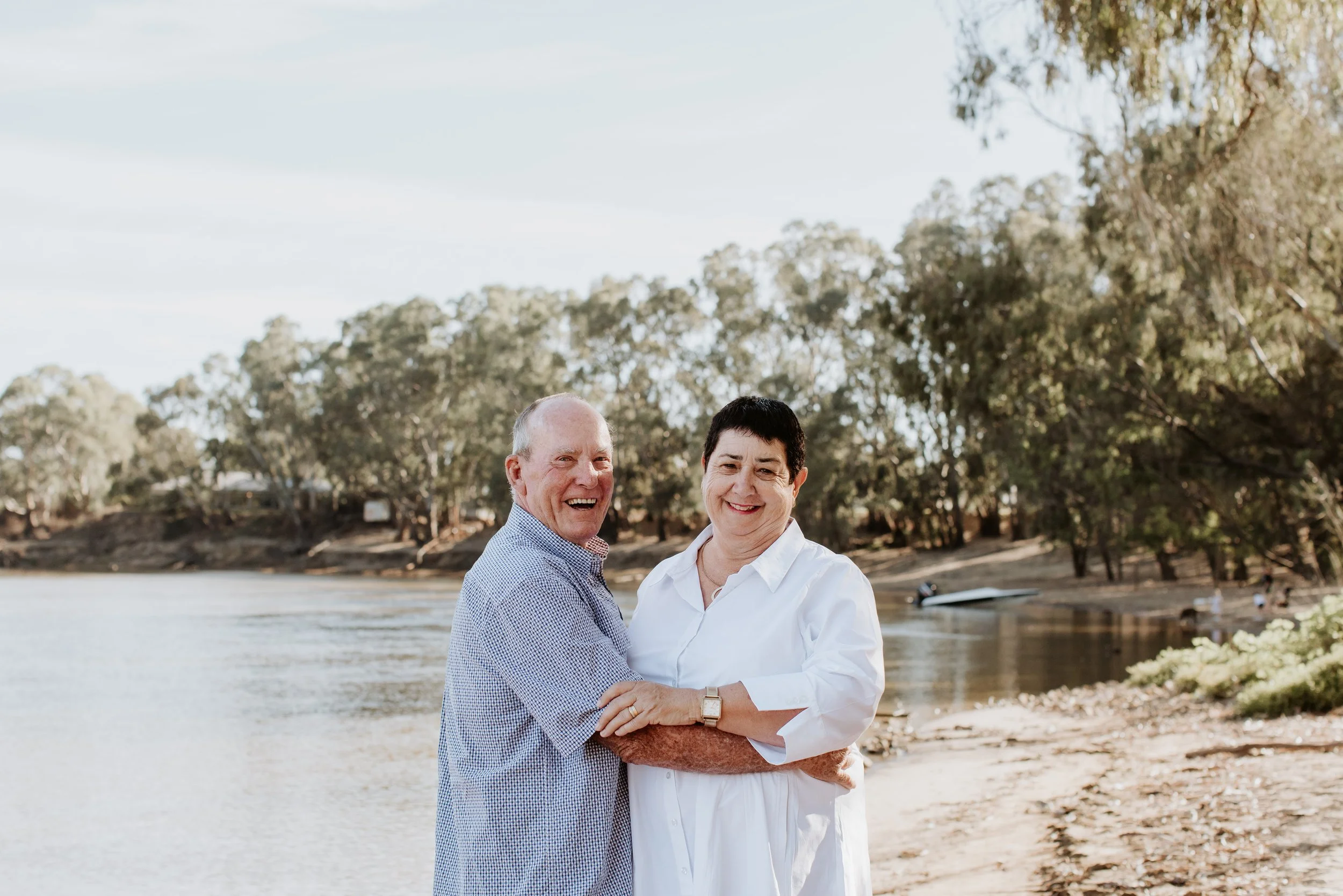 A happy older man and woman embracing each other on a sandy riverbank with trees in the background.