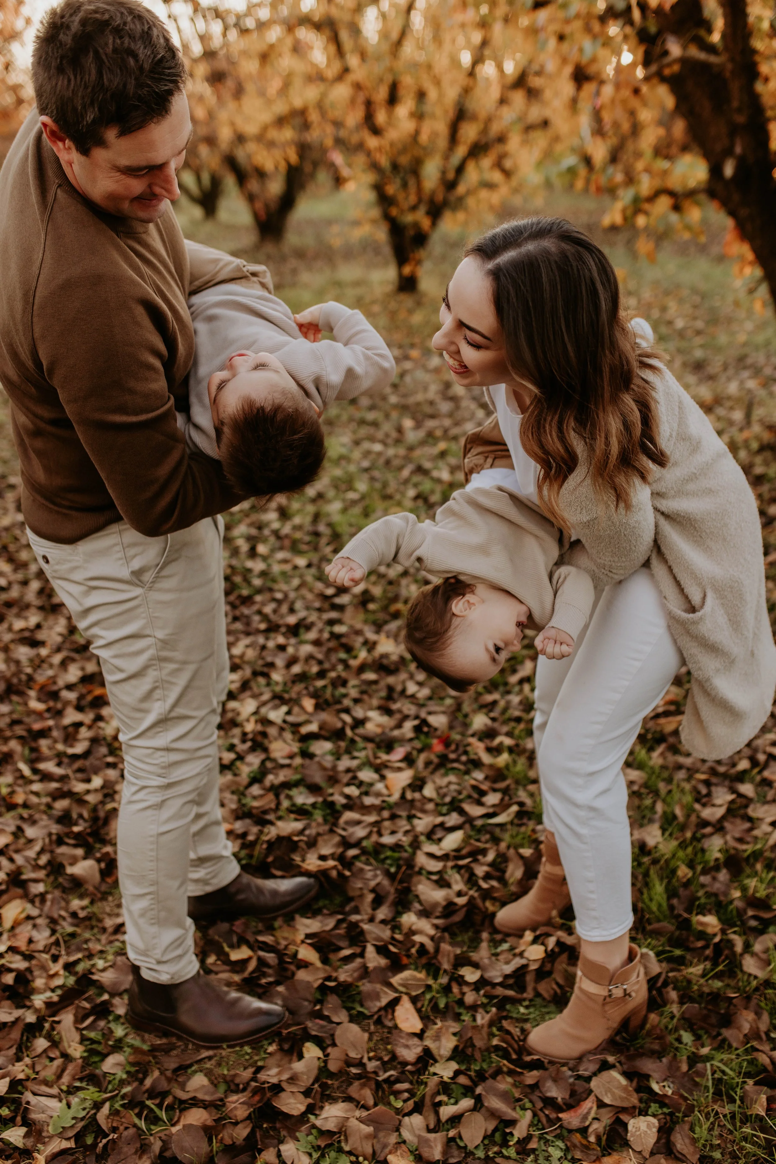 A family of four playing and laughing in an autumn forest, with fall leaves on the ground. The father is holding a young boy upside down, and the mother is helping a girl upside down, all smiling and having fun.