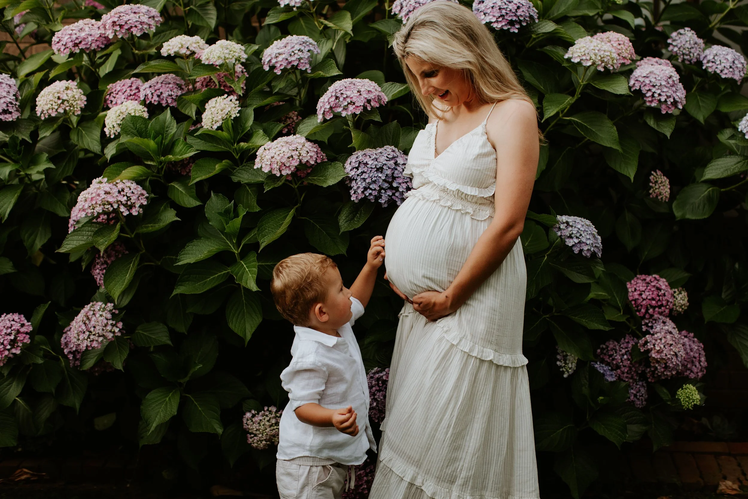 A pregnant woman smiling and holding her belly, standing next to a young boy touching her belly, in front of blooming pink and purple hydrangea flowers.
